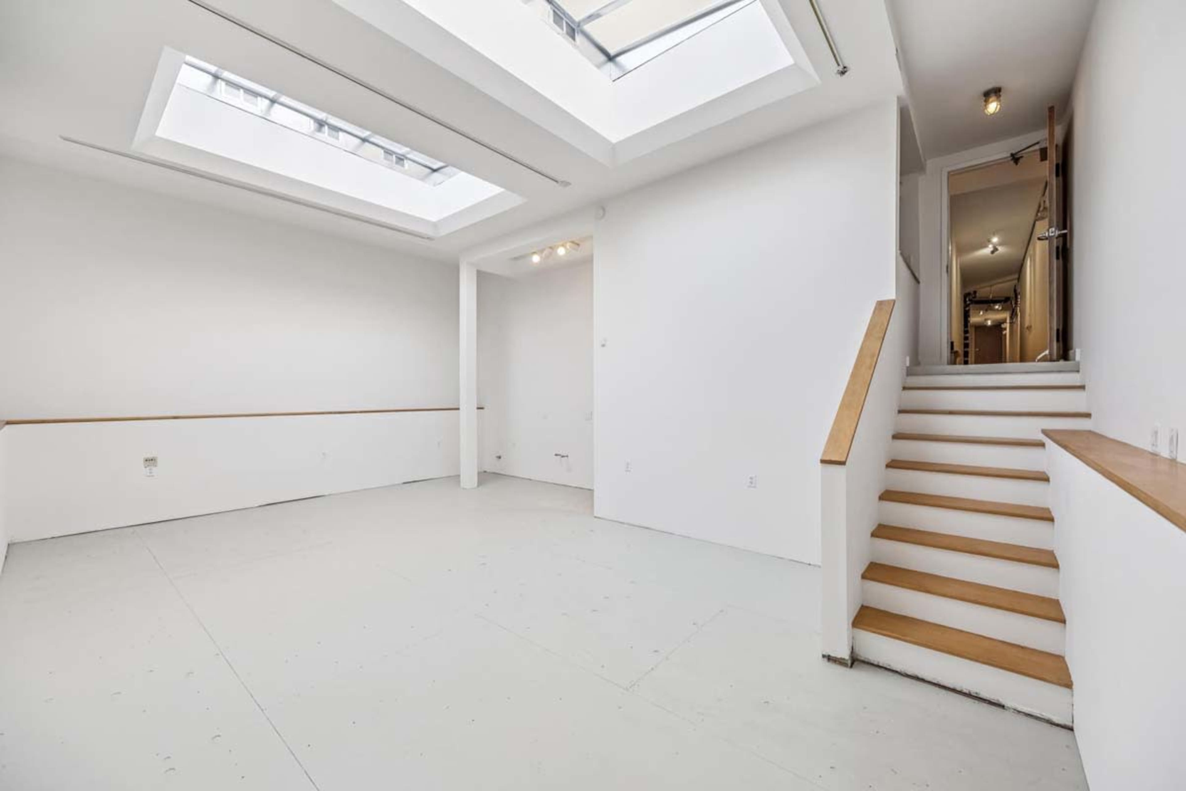 The image shows an empty, well-lit room with high ceilings, a staircase on one side, and a skylight above.