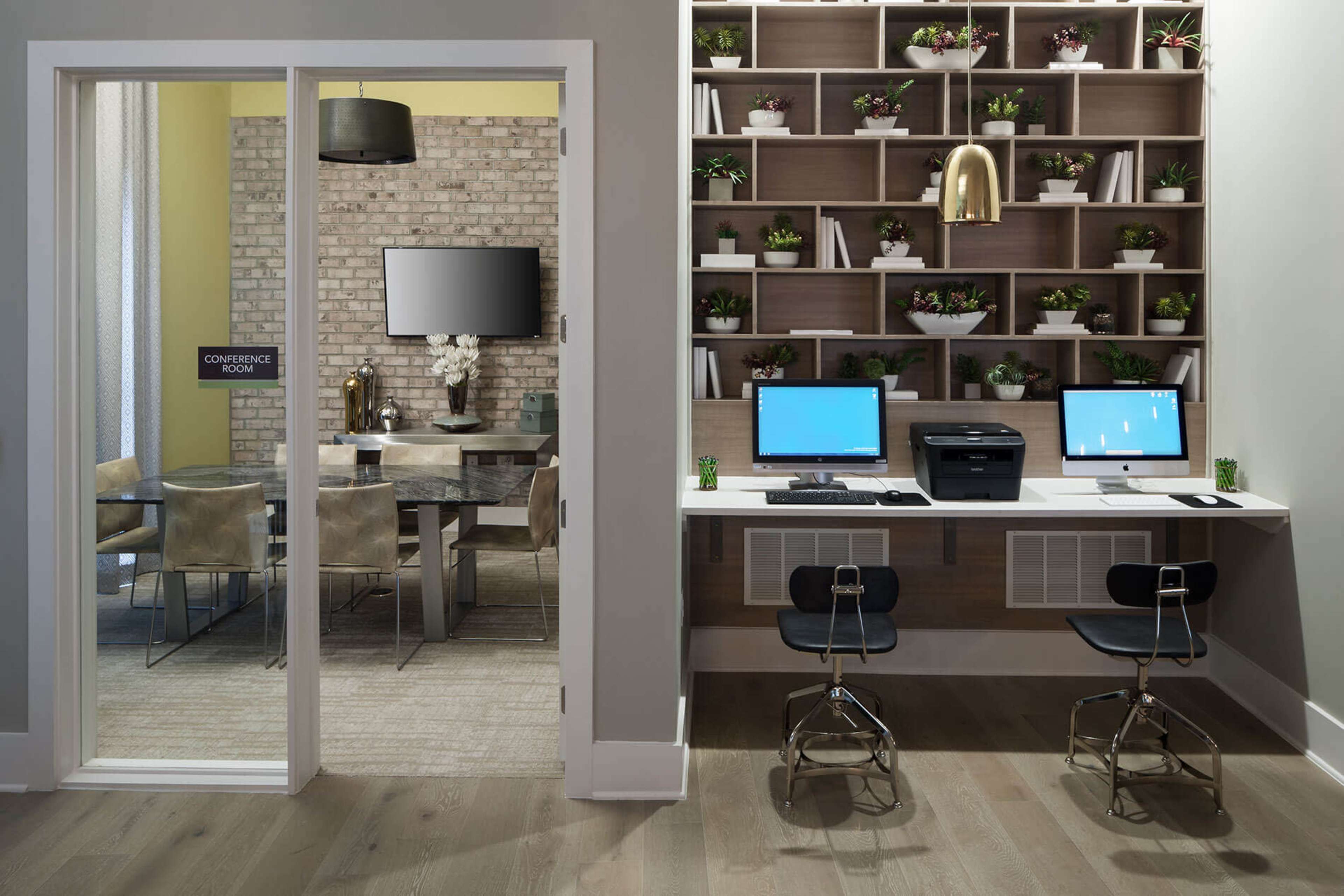 A modern office space featuring a conference room with a large table and a separate work area with two computers and a printer against a wall of shelves filled with plants.