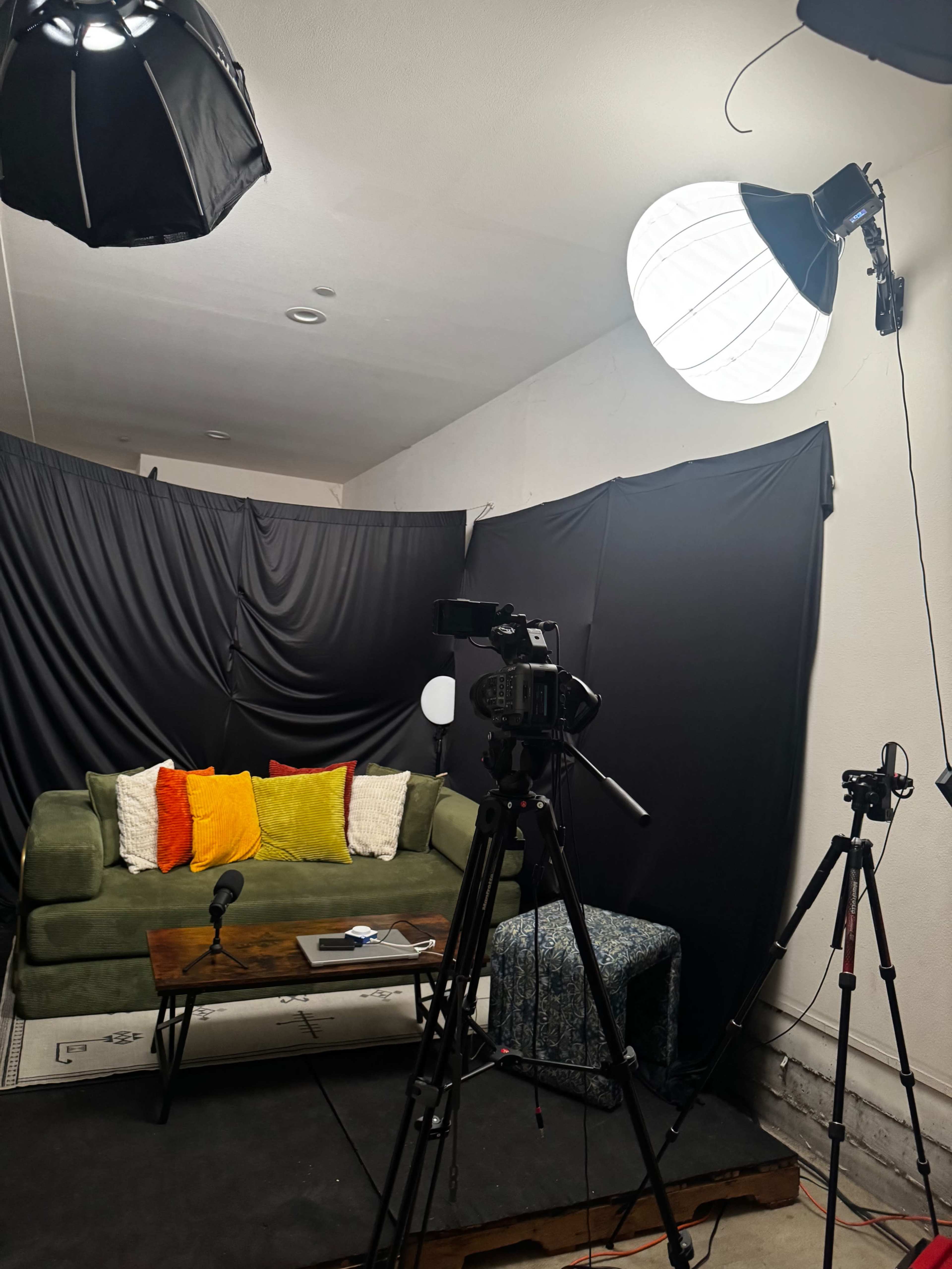 The scene shows a small studio setup with a green sofa adorned with colorful pillows, a wooden table with a device on it, and two cameras positioned for filming, surrounded by black backdrops and lighting equipment.