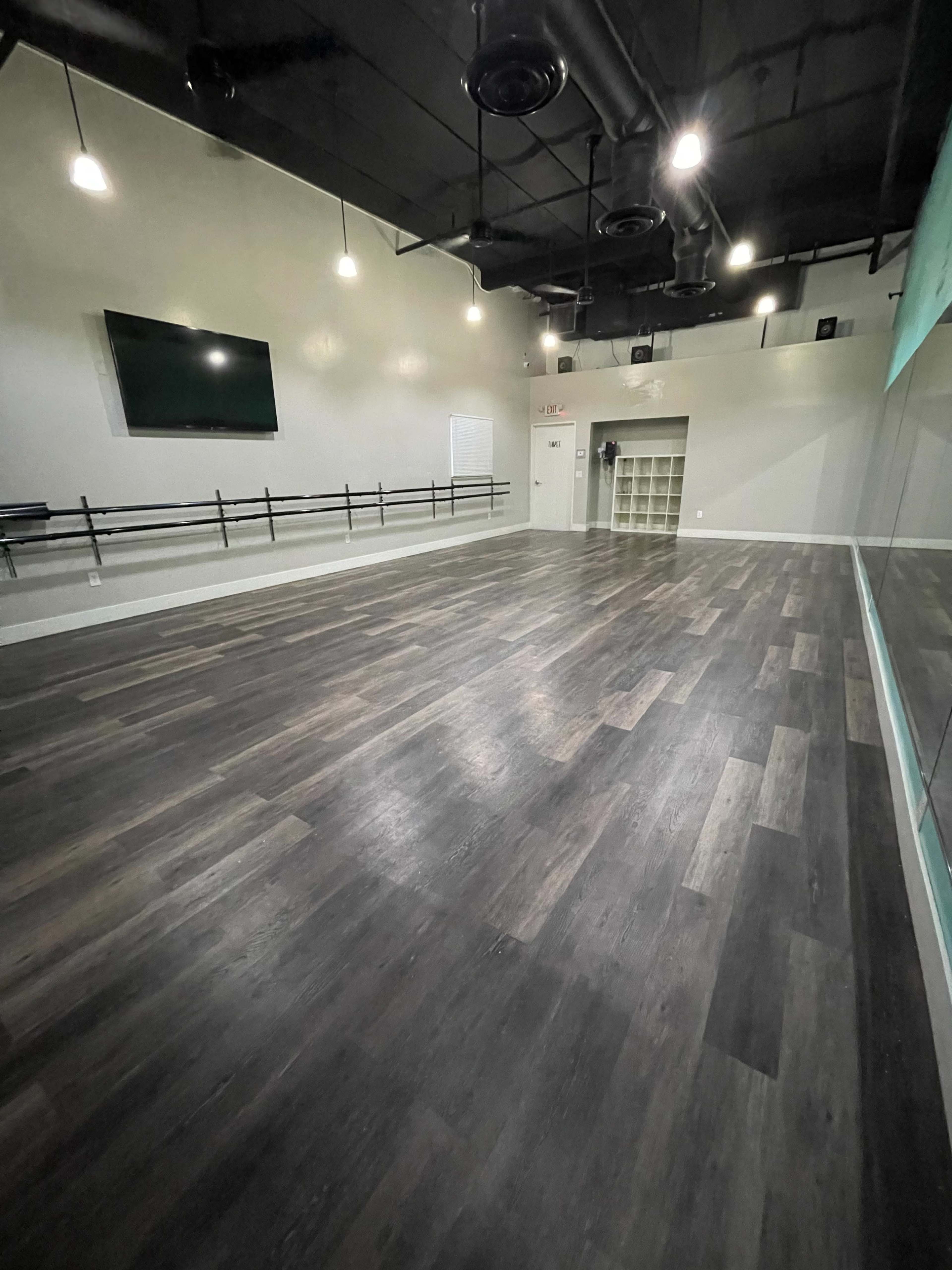 A spacious dance studio with dark wood flooring, mirrored walls, and a large wall-mounted screen.