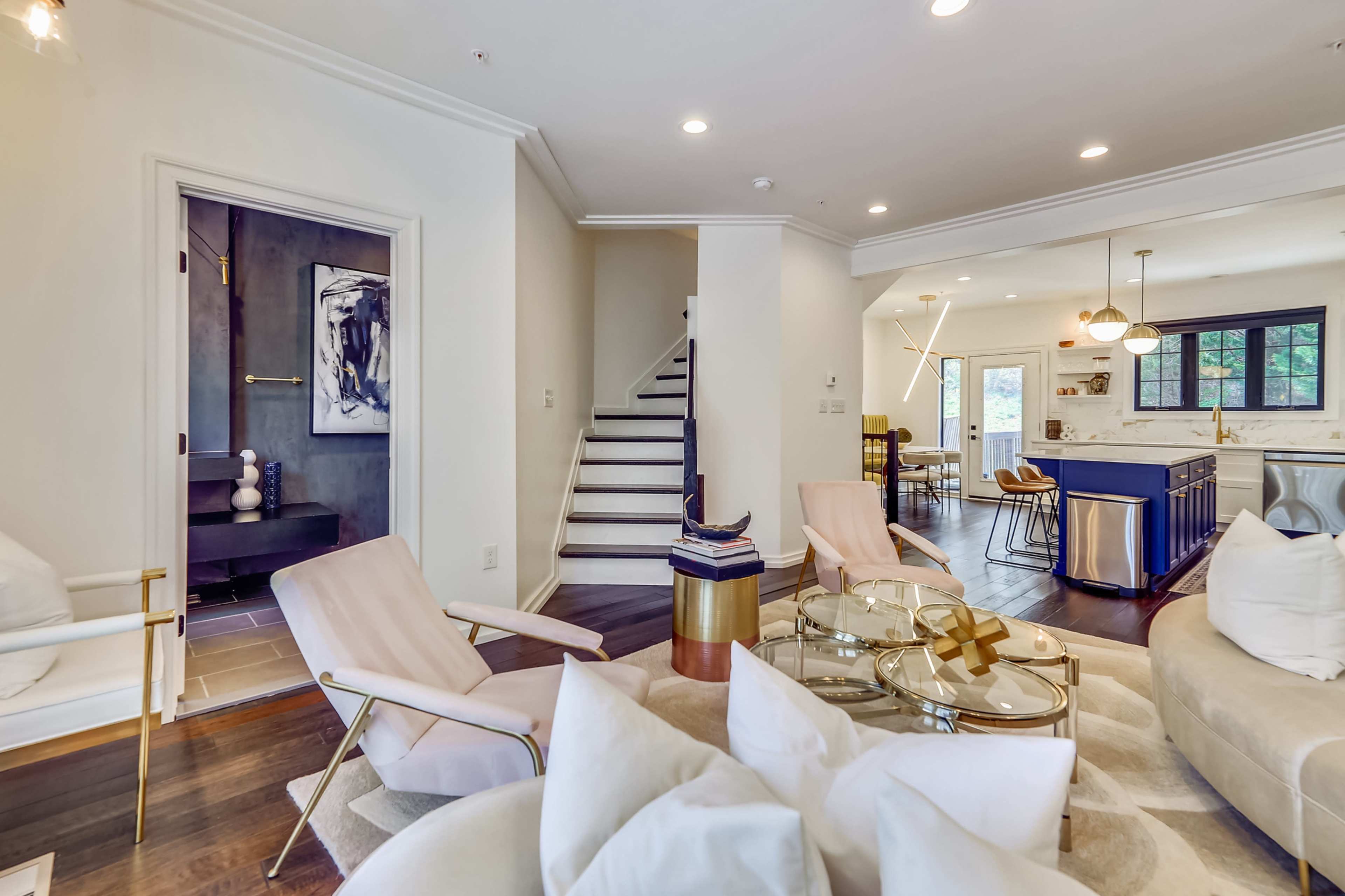 The image shows a well-lit interior space featuring a modern living room with a round glass coffee table, two pink chairs, and a staircase leading to an upper level.