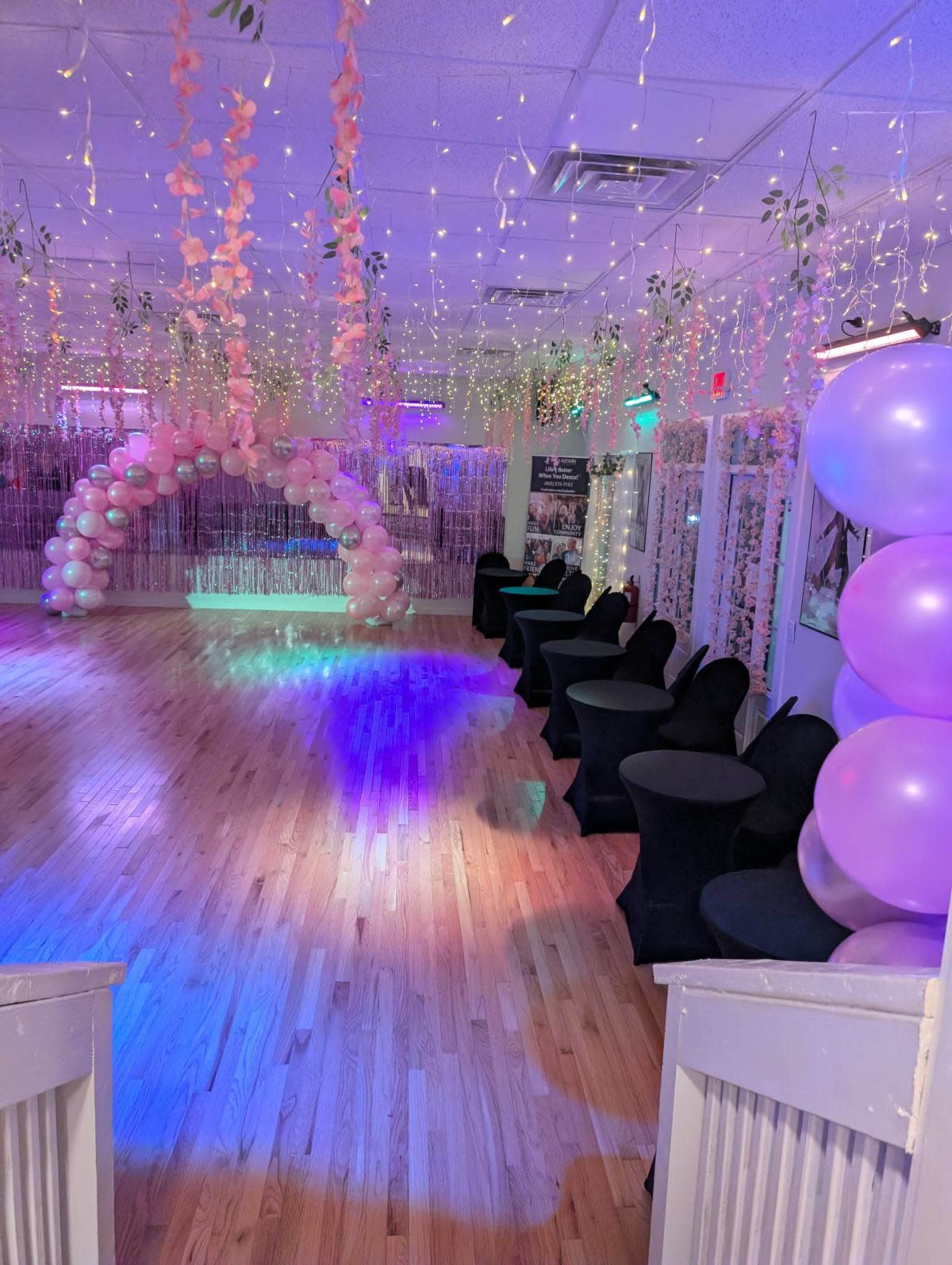 The image shows a party venue decorated with pink and purple balloons, draped lights, and floral arrangements, featuring a wooden floor and black chairs arranged along the sides.