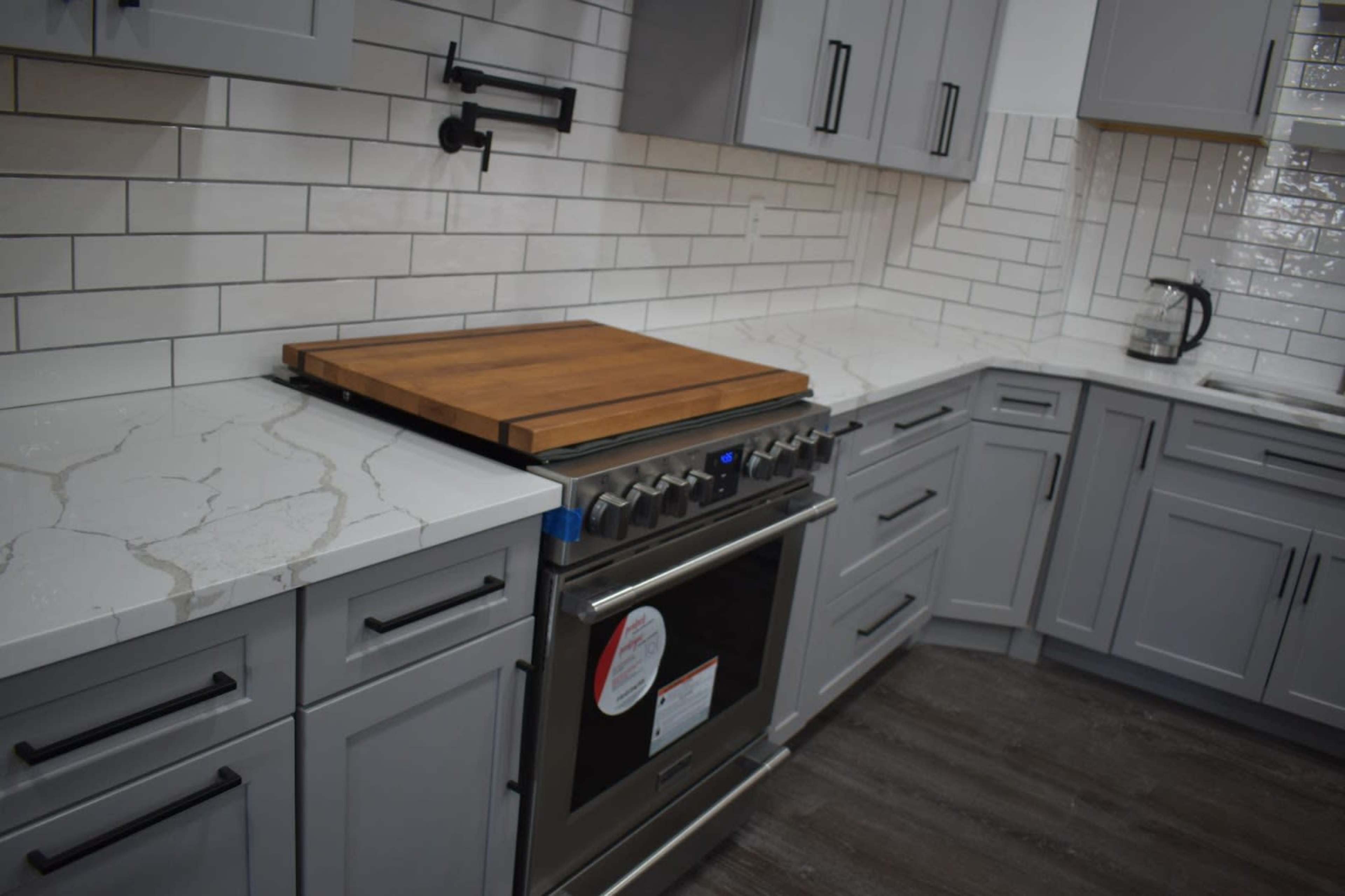 A modern kitchen with gray cabinets, a stainless steel range, a wooden cutting board on the countertop, and white tiled backsplash.