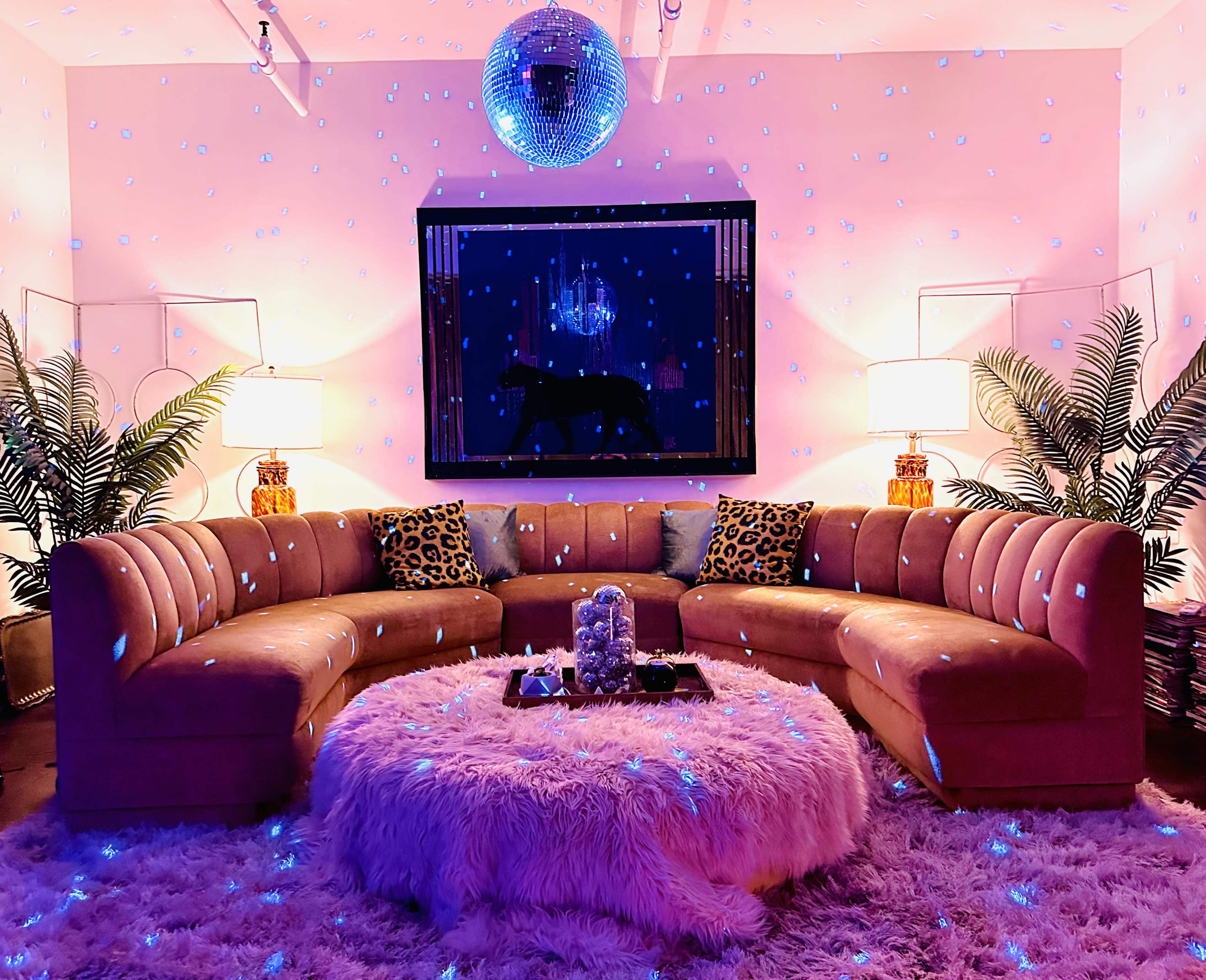A spacious living room features a curved velvet sofa, a round shag rug, and disco ball lighting, with palm plants and framed artwork on the walls.