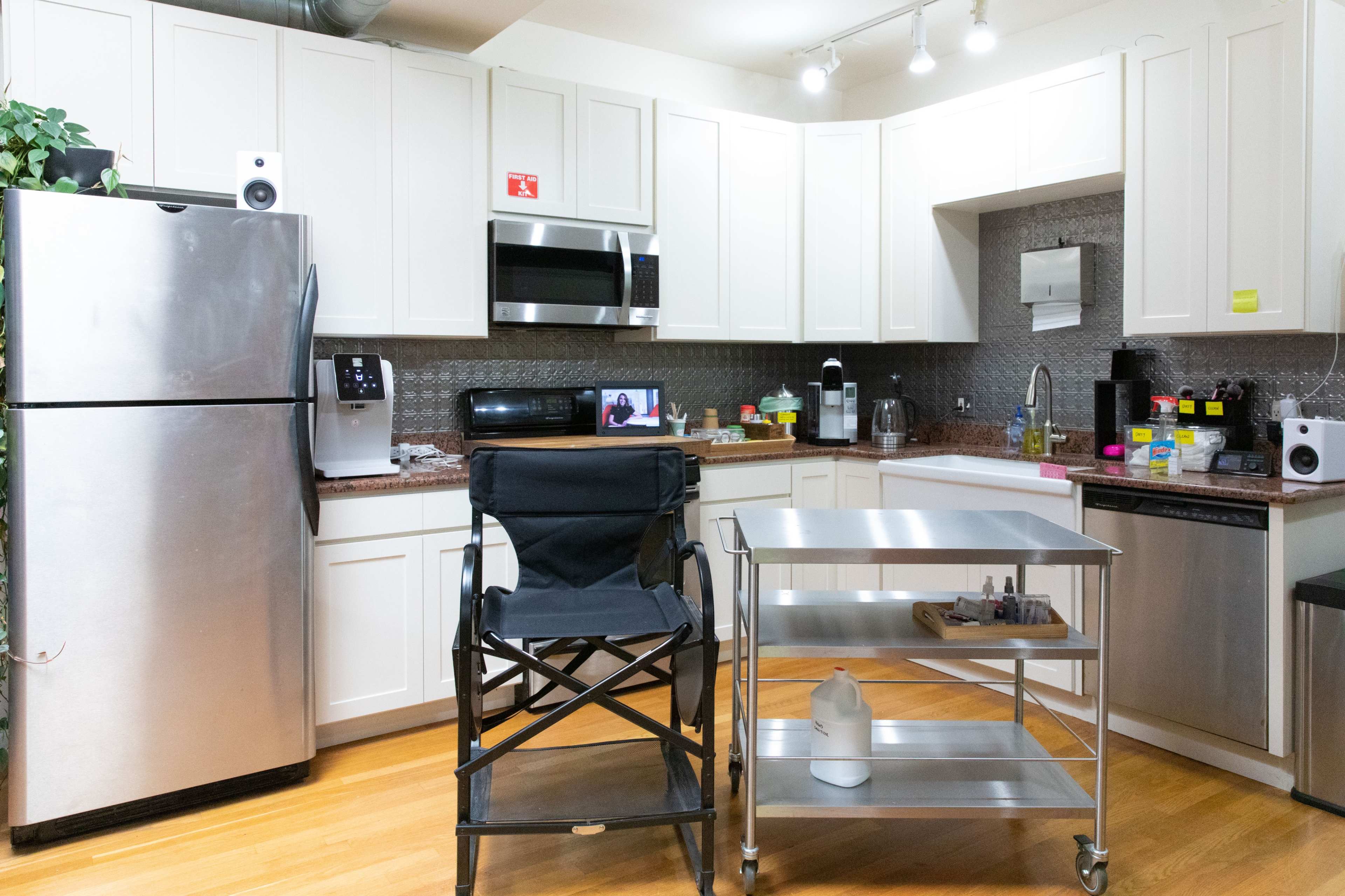 The image shows a modern kitchen with white cabinets, stainless steel appliances, and a folding chair next to a metal serving cart.