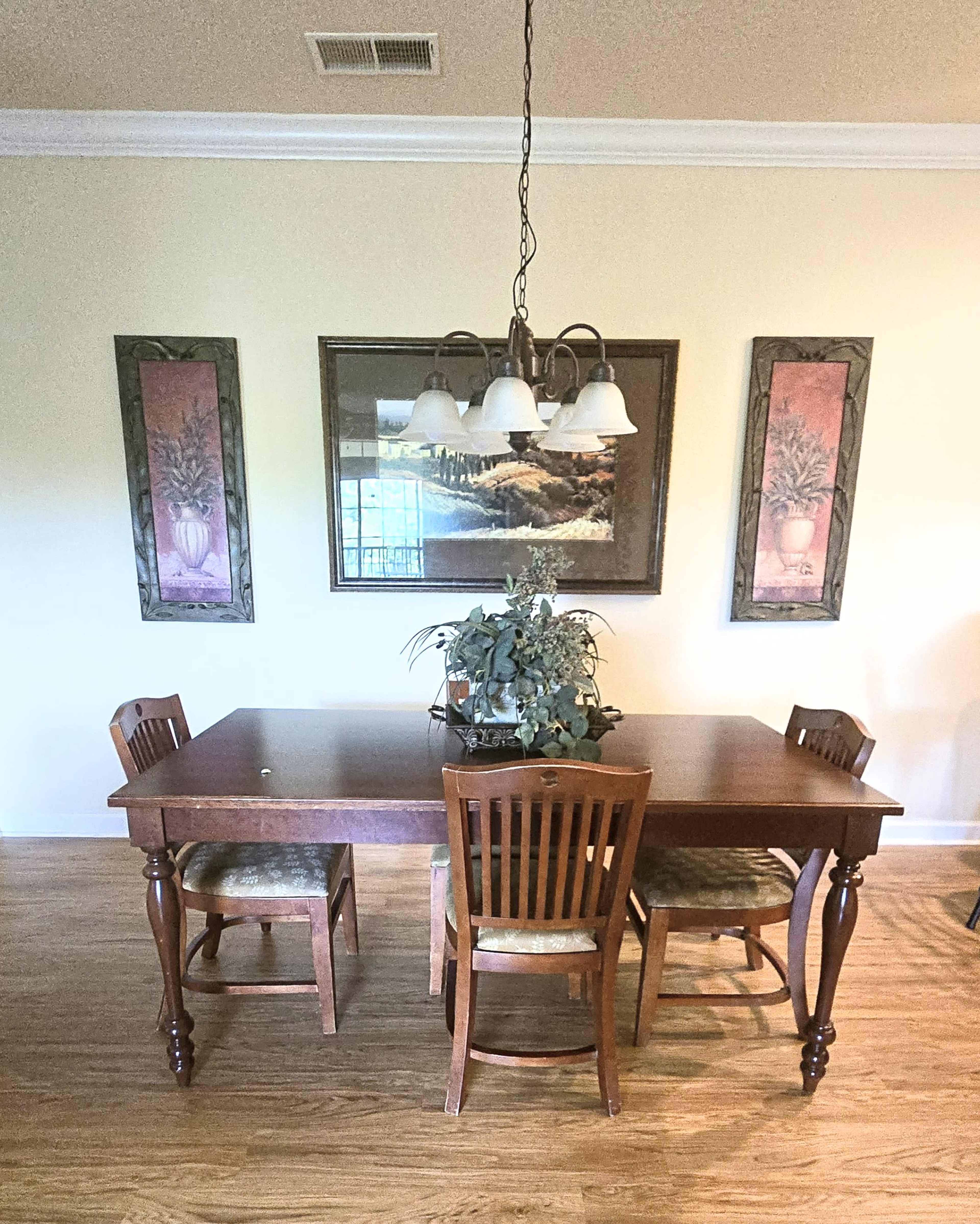 A wooden dining table with three chairs and a plant centerpiece is positioned under a chandelier in a room adorned with framed artwork.