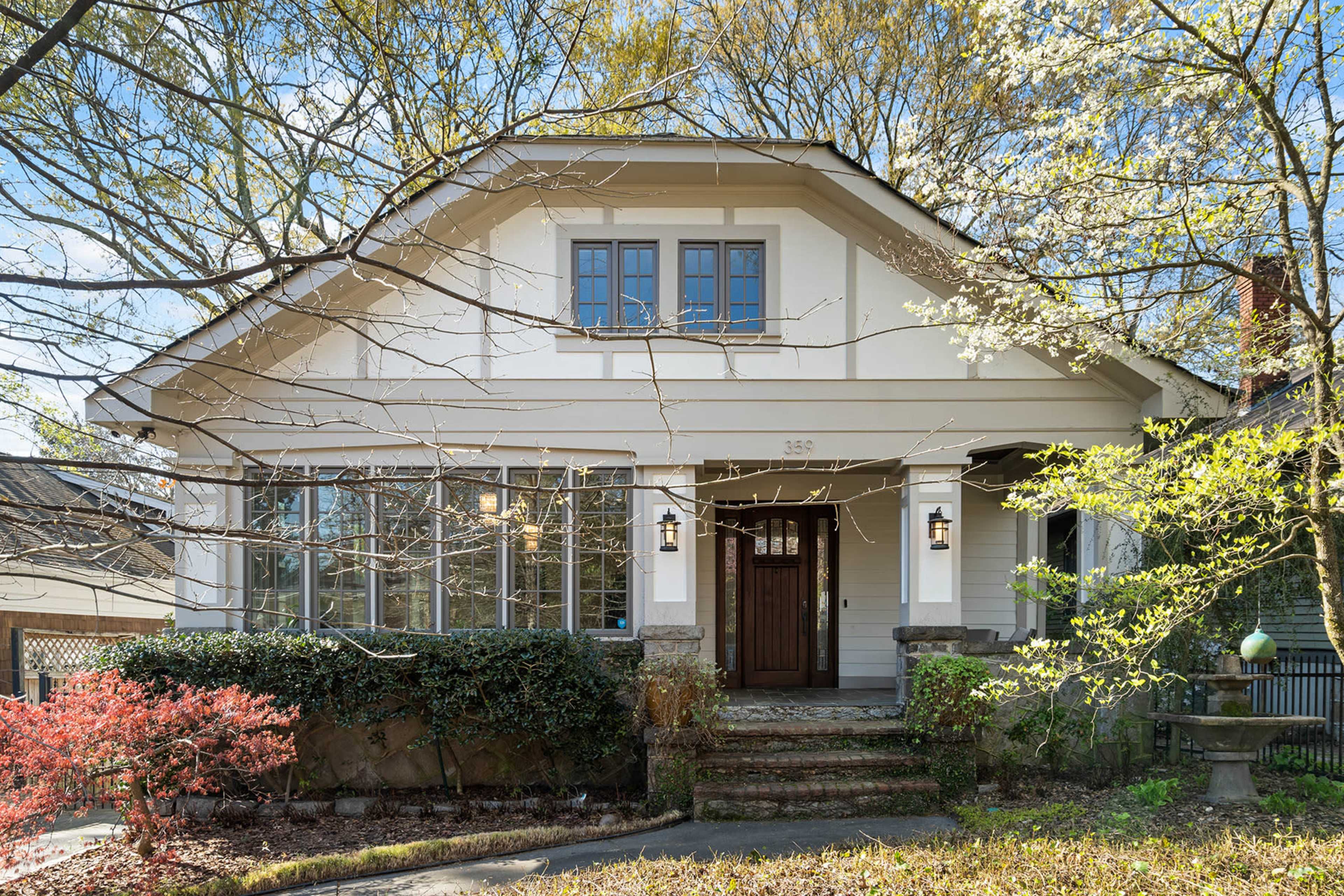 A bungalow-style house with a covered porch and stone steps is surrounded by trees and shrubs.