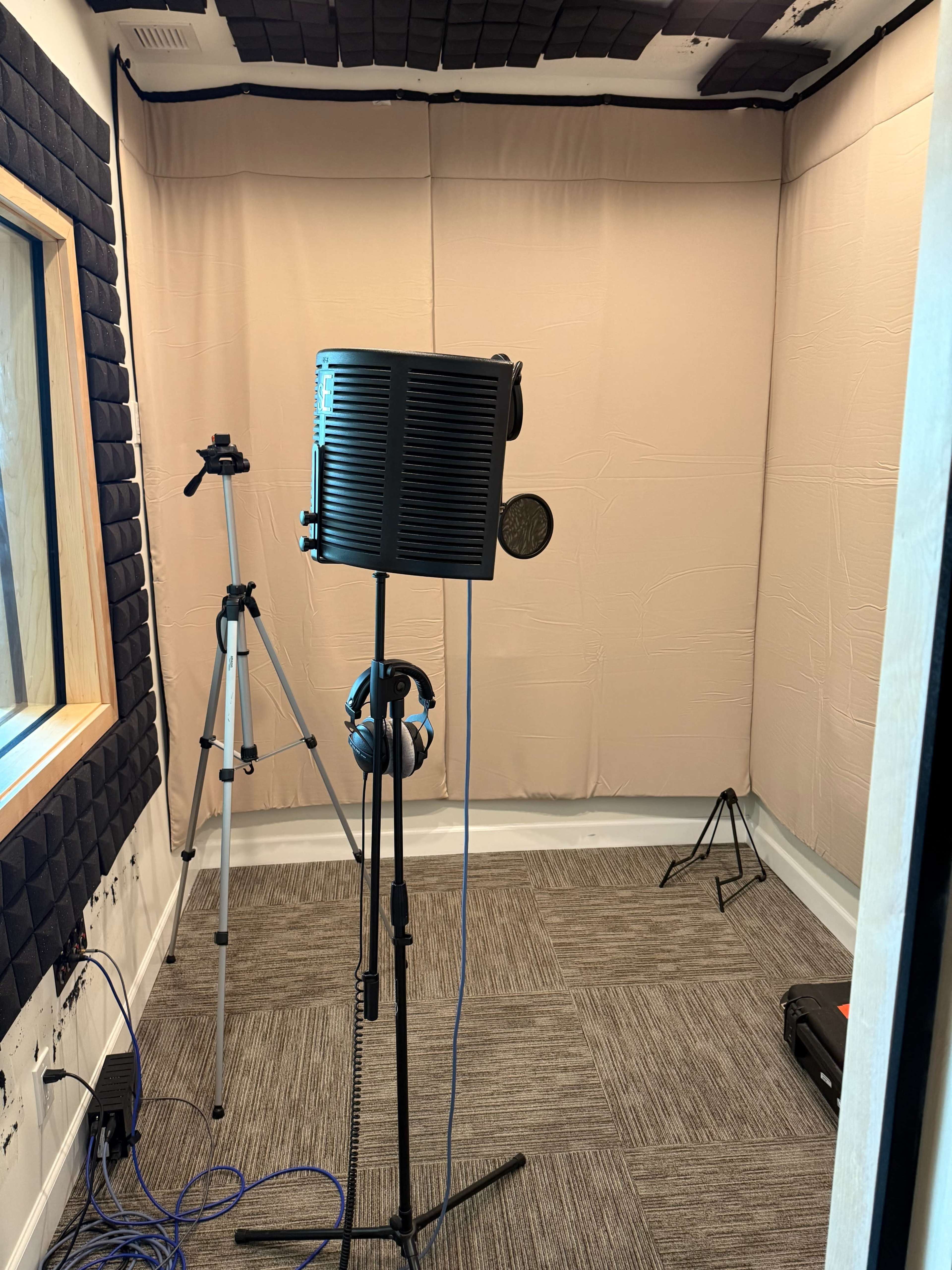 The image shows a soundproof recording studio with a microphone on a stand, headphones, a camera tripod, and sound-absorbing panels on the walls.