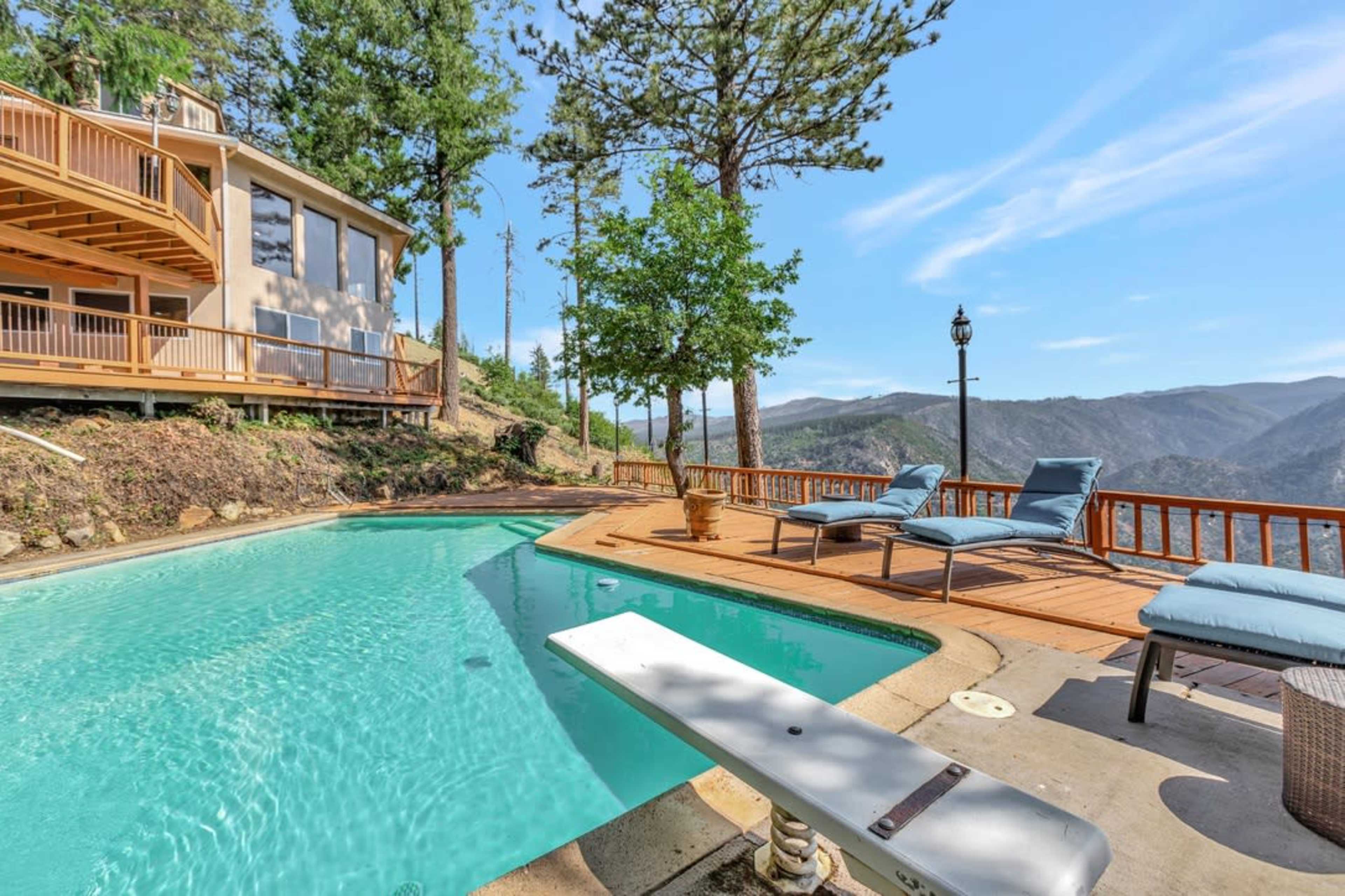 A clear swimming pool sits beside a wooden deck, surrounded by trees and overlooking a mountainous landscape.