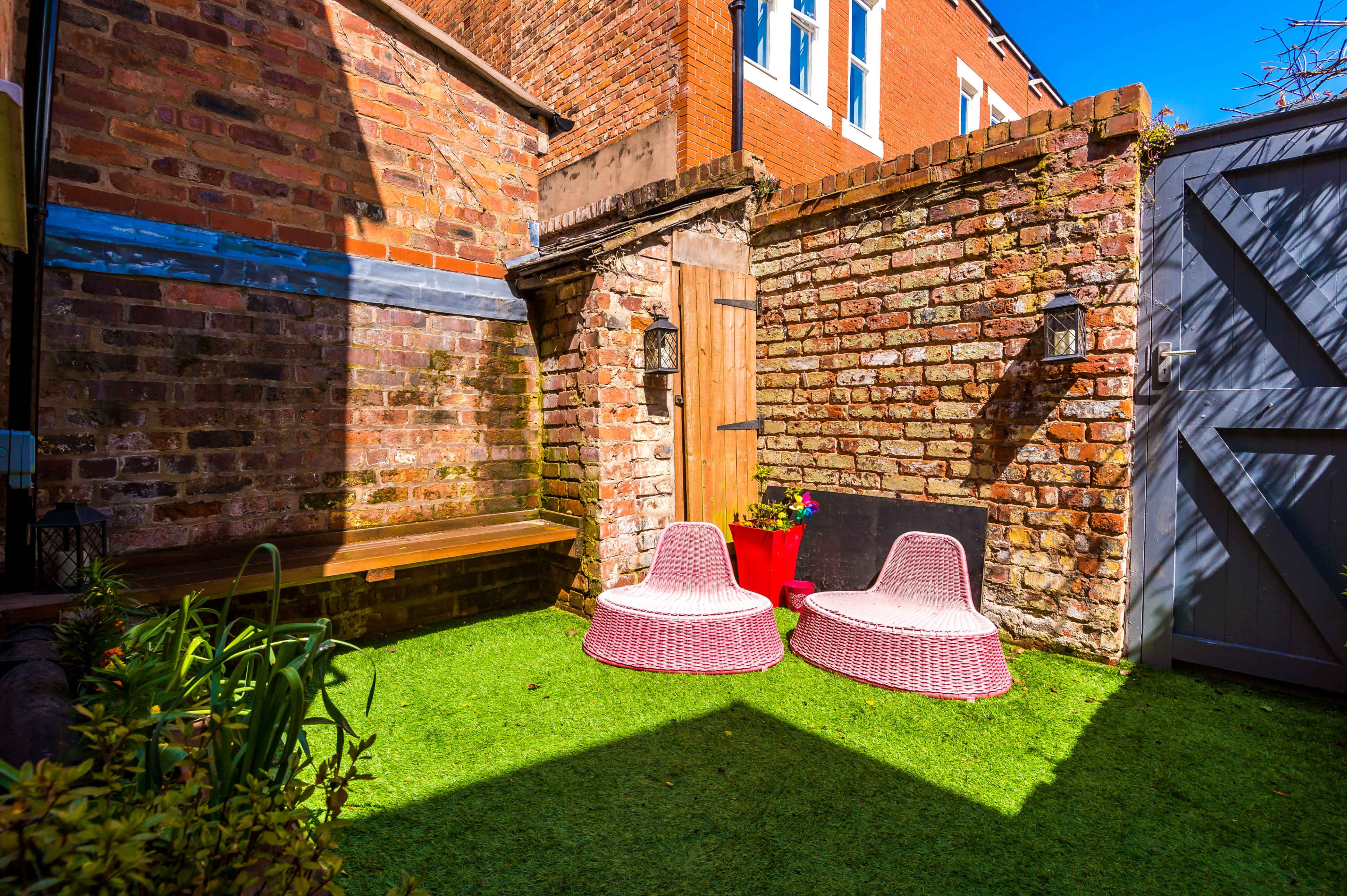 A small courtyard with two pink, wicker chairs on artificial grass, surrounded by brick walls and a wooden door.