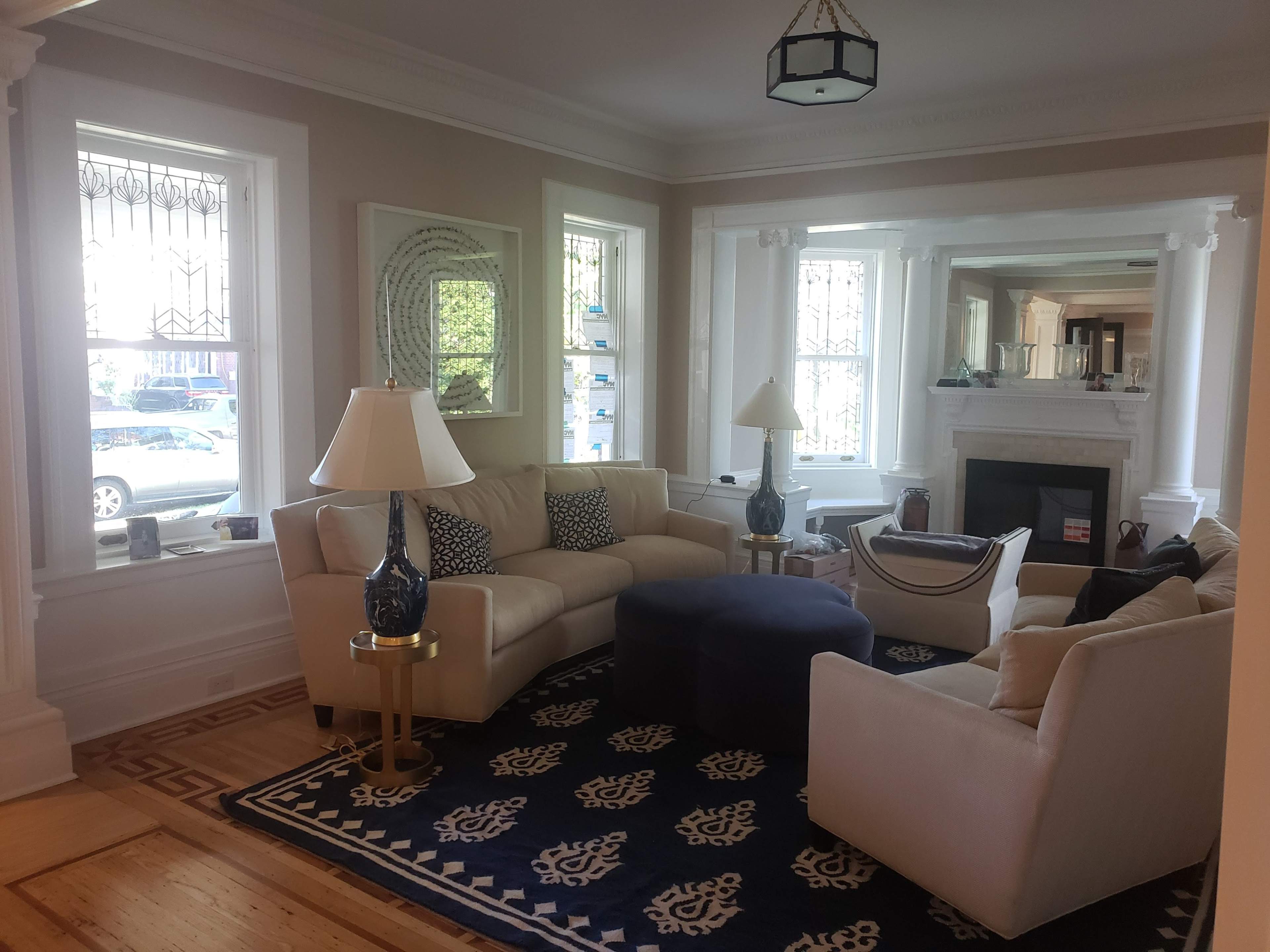 The image depicts a neatly arranged living room with two couches, a round ottoman, and a coffee table, surrounded by large windows and a decorative rug.