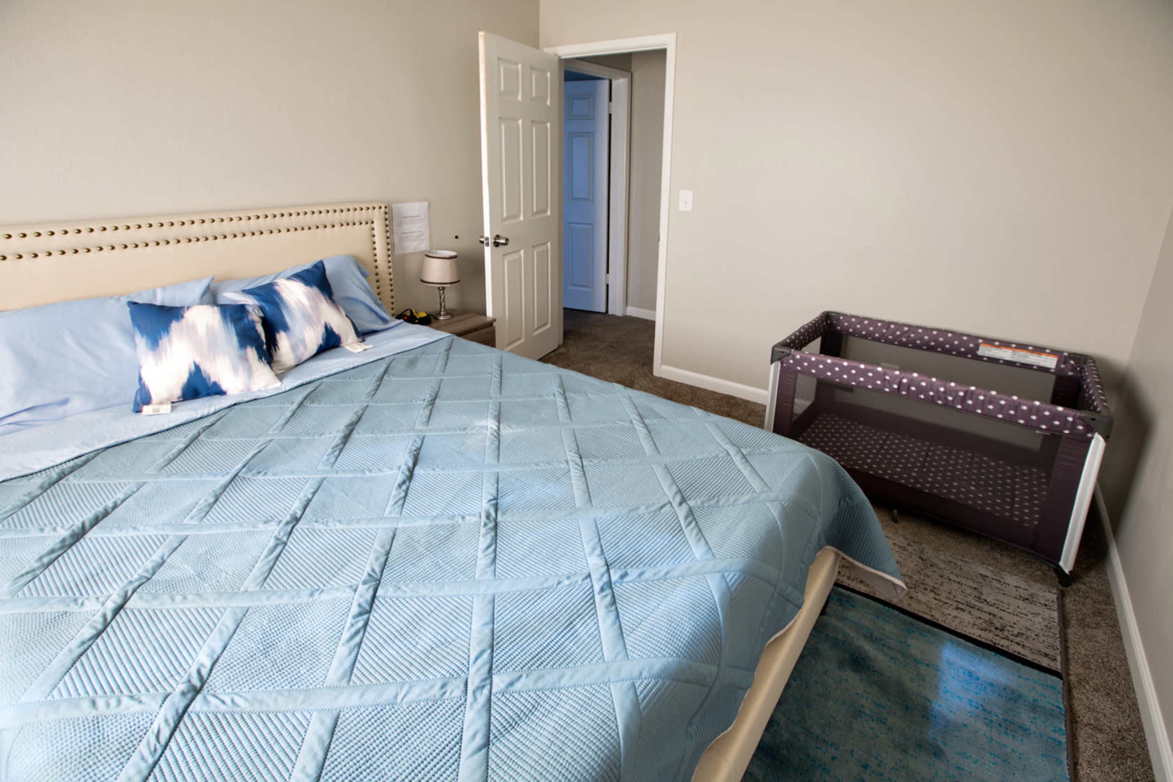 A bedroom with a queen-sized bed covered in a blue quilt and a portable crib in the corner, with a door leading to another room visible in the background.