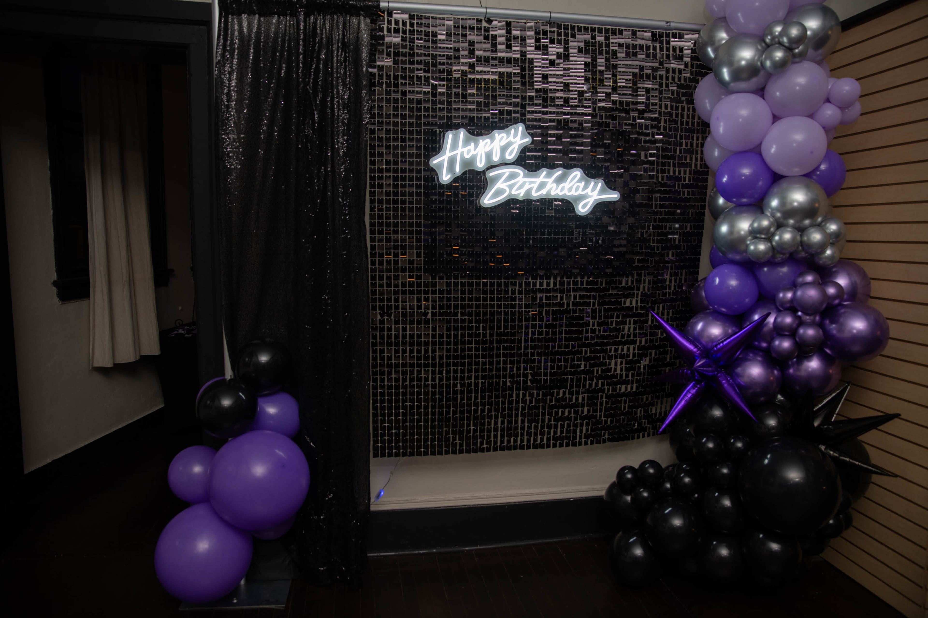 A birthday decoration setup features a neon "Happy Birthday" sign against a mirrored backdrop, surrounded by black and purple balloons.