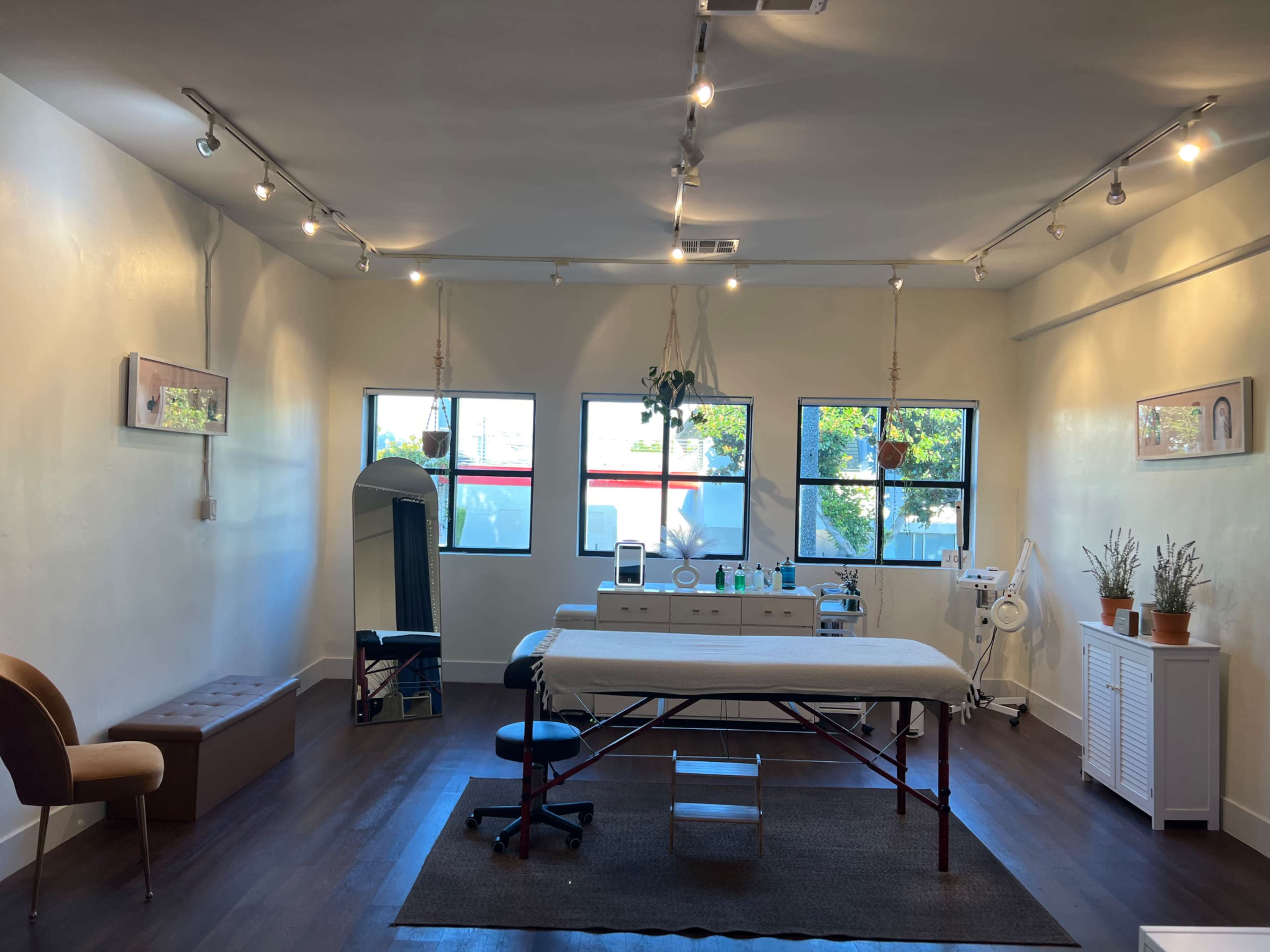 The image shows a bright, clean treatment room with a massage table, large windows, and various equipment arranged along the walls.