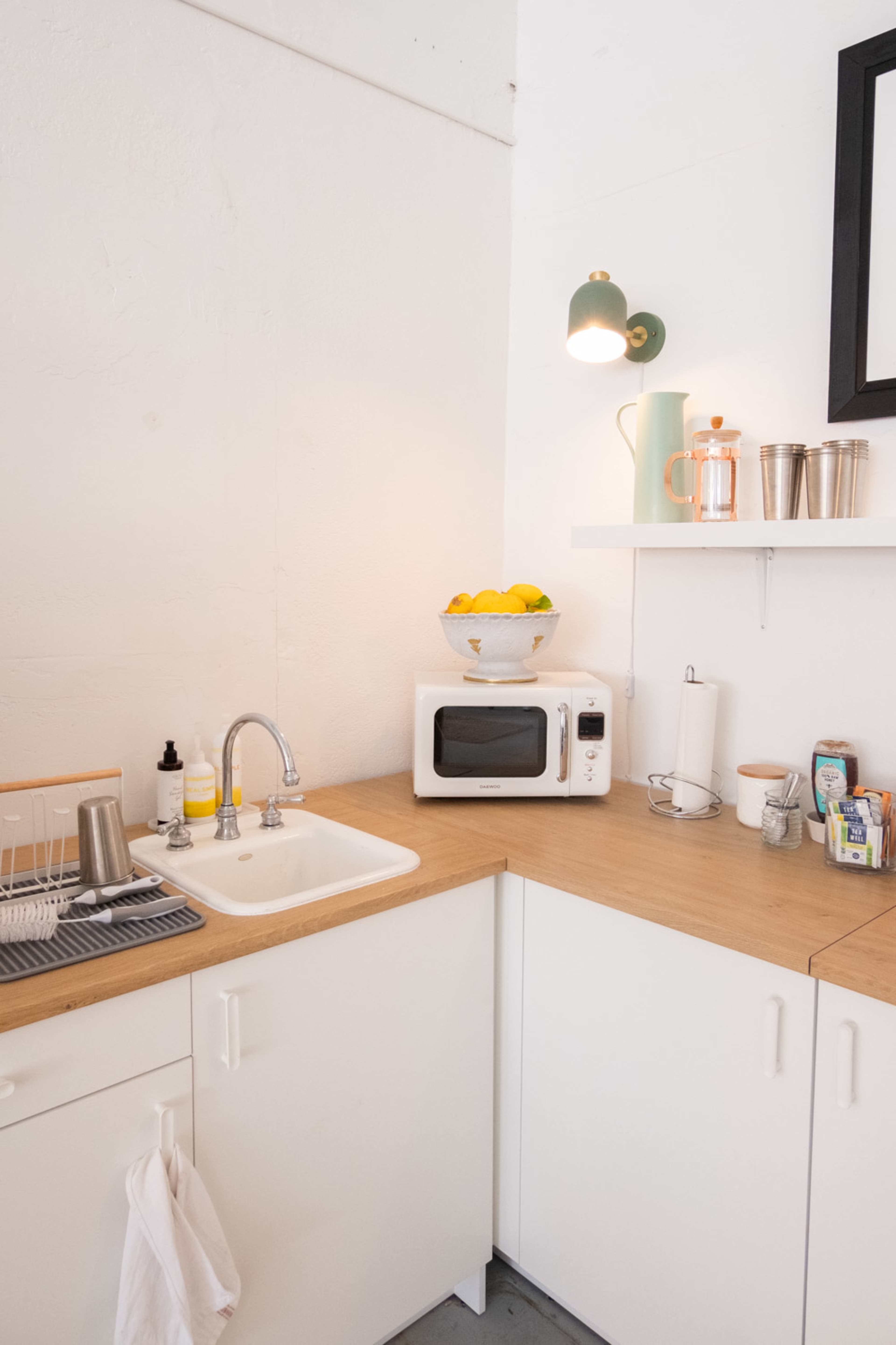 A corner kitchen features a white sink, a microwave on a countertop, a bowl of lemons above, and neatly arranged kitchen items on a shelf.