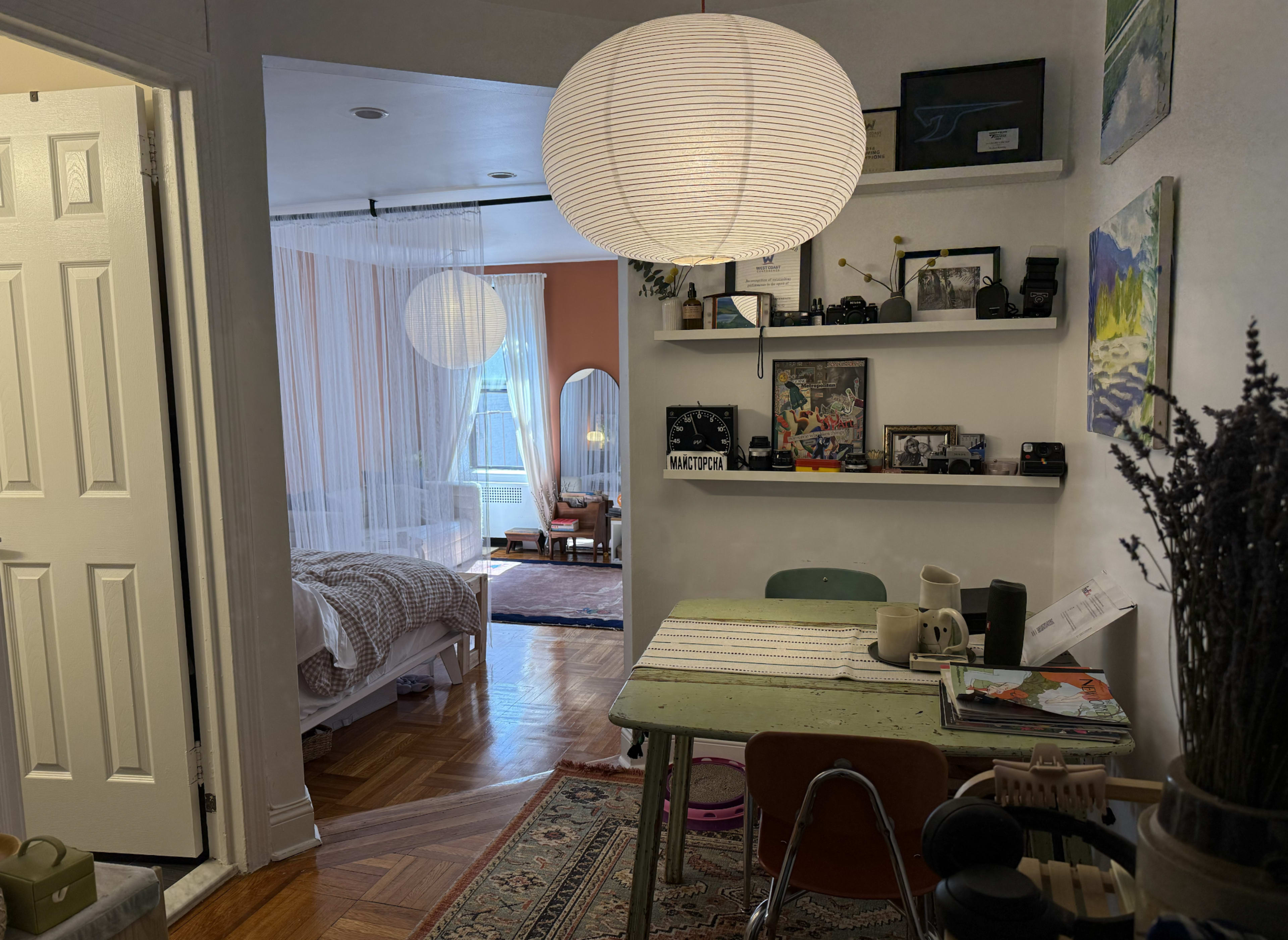 The image shows a cozy, well-lit apartment with a dining area featuring a wooden table and chairs, leading into a bedroom area with a large window and soft curtains.