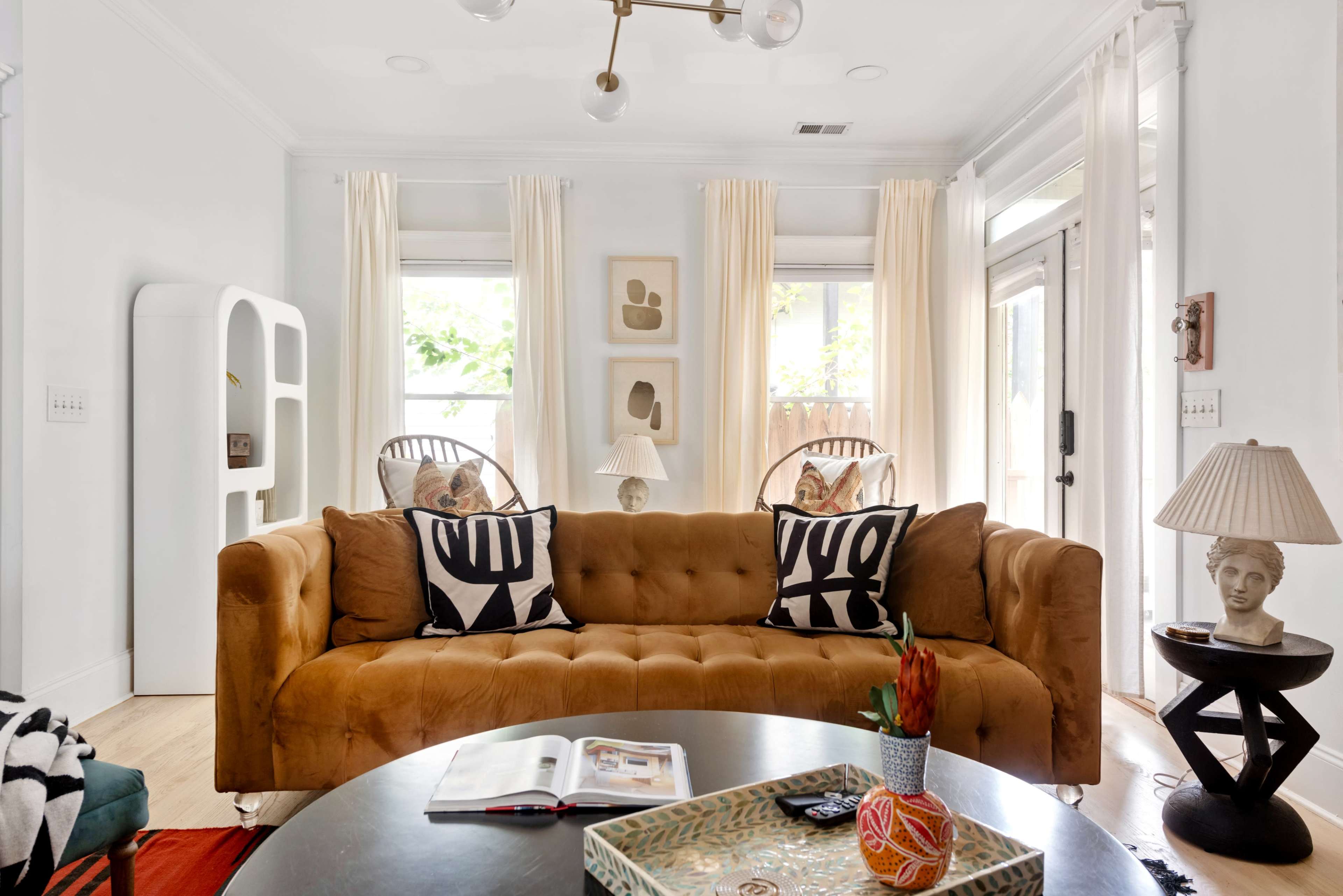 The room features a cozy sofa with patterned cushions, a round coffee table, and large windows with light curtains, creating a bright and inviting living space.