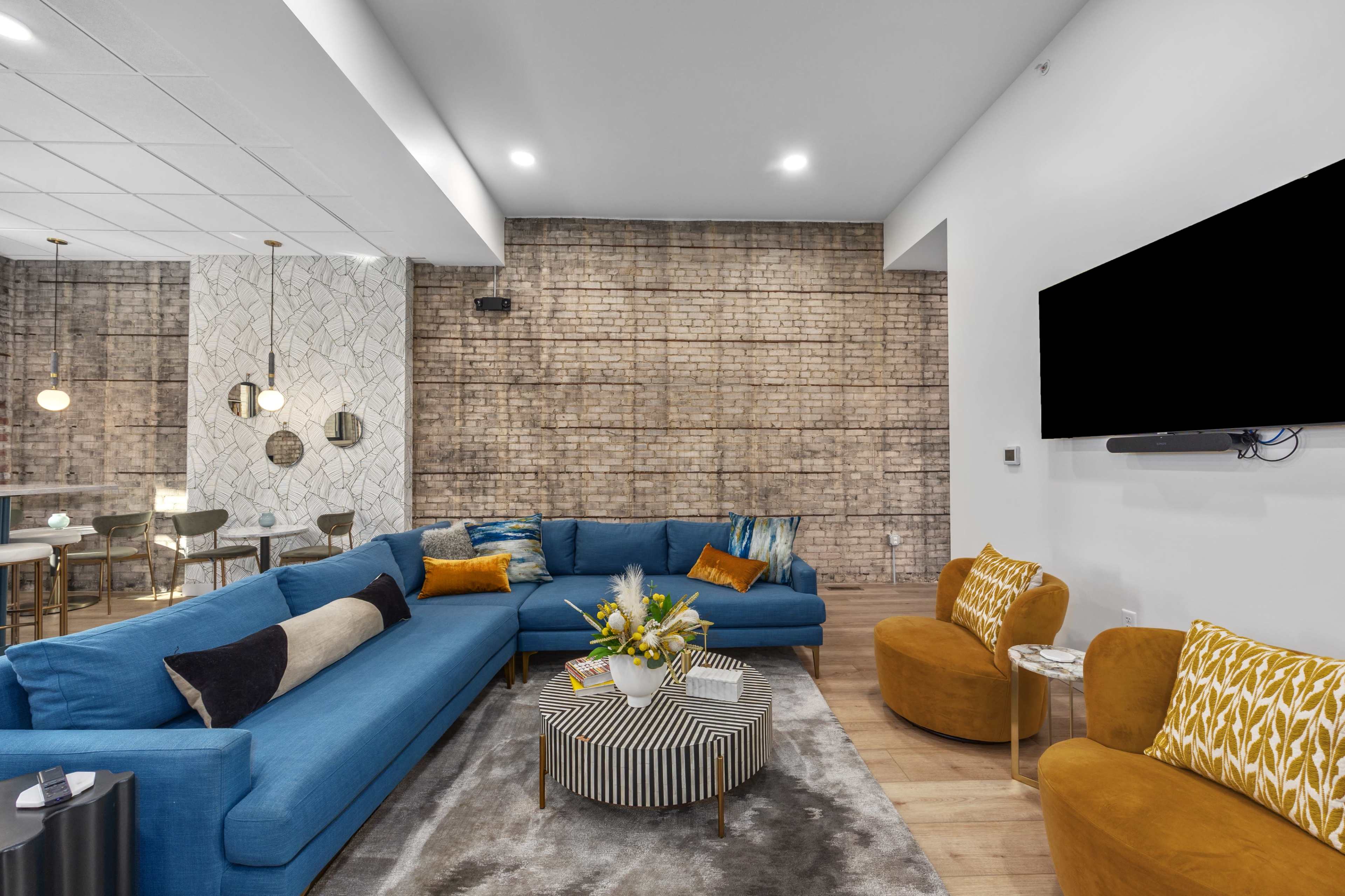 A modern living room features a blue sectional sofa, a striped coffee table, and orange accent chairs, set against a textured brick wall.