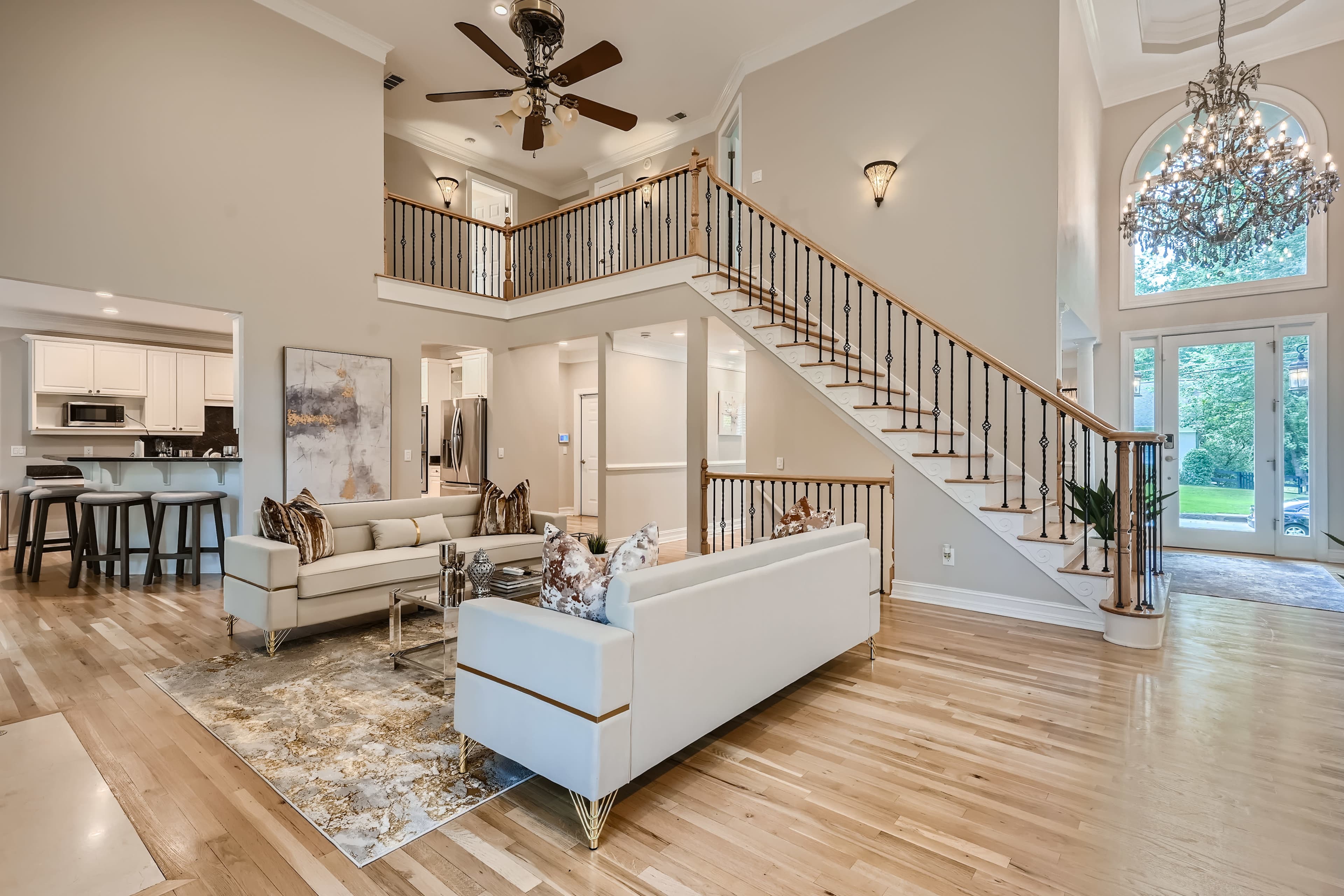 A spacious living room with a large staircase, elegant furniture, and a high ceiling adorned with a chandelier, all set in a well-lit, open space.