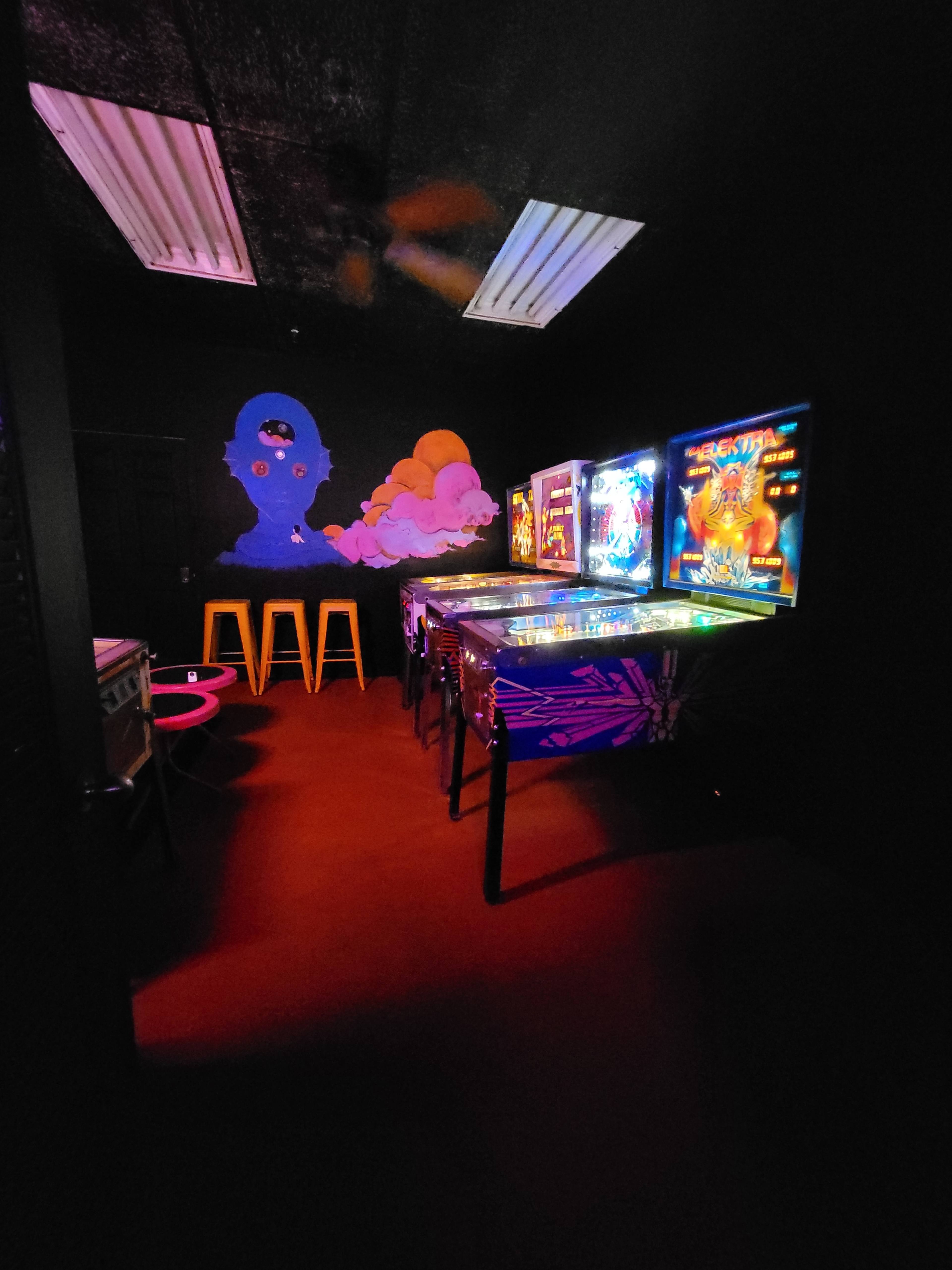 A dimly lit room features several pinball machines and colorful stools against a black wall with a mural.