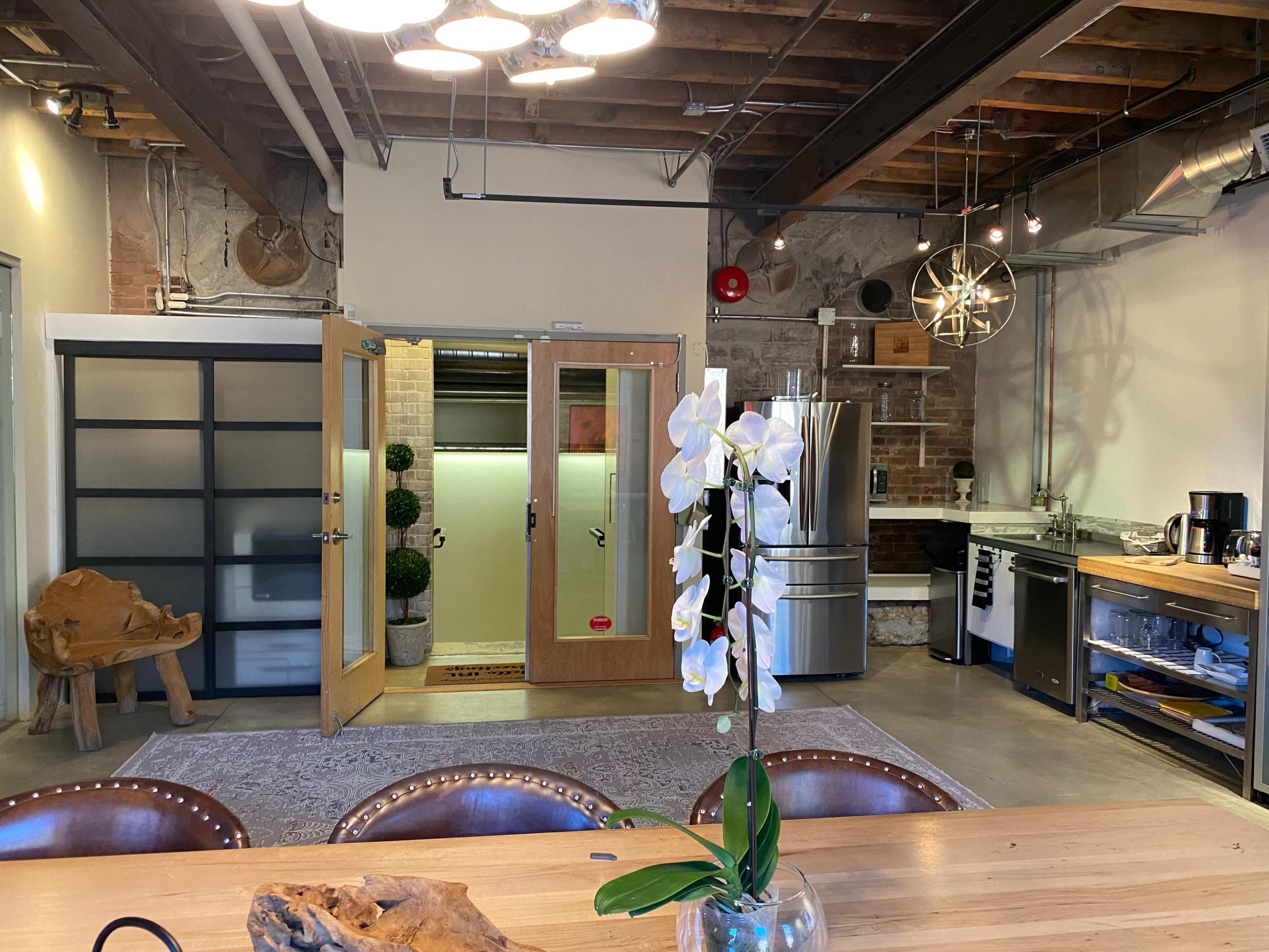 A modern kitchen and dining area with a wooden table, flowering plant, metal appliances, and a door leading to a separate room.