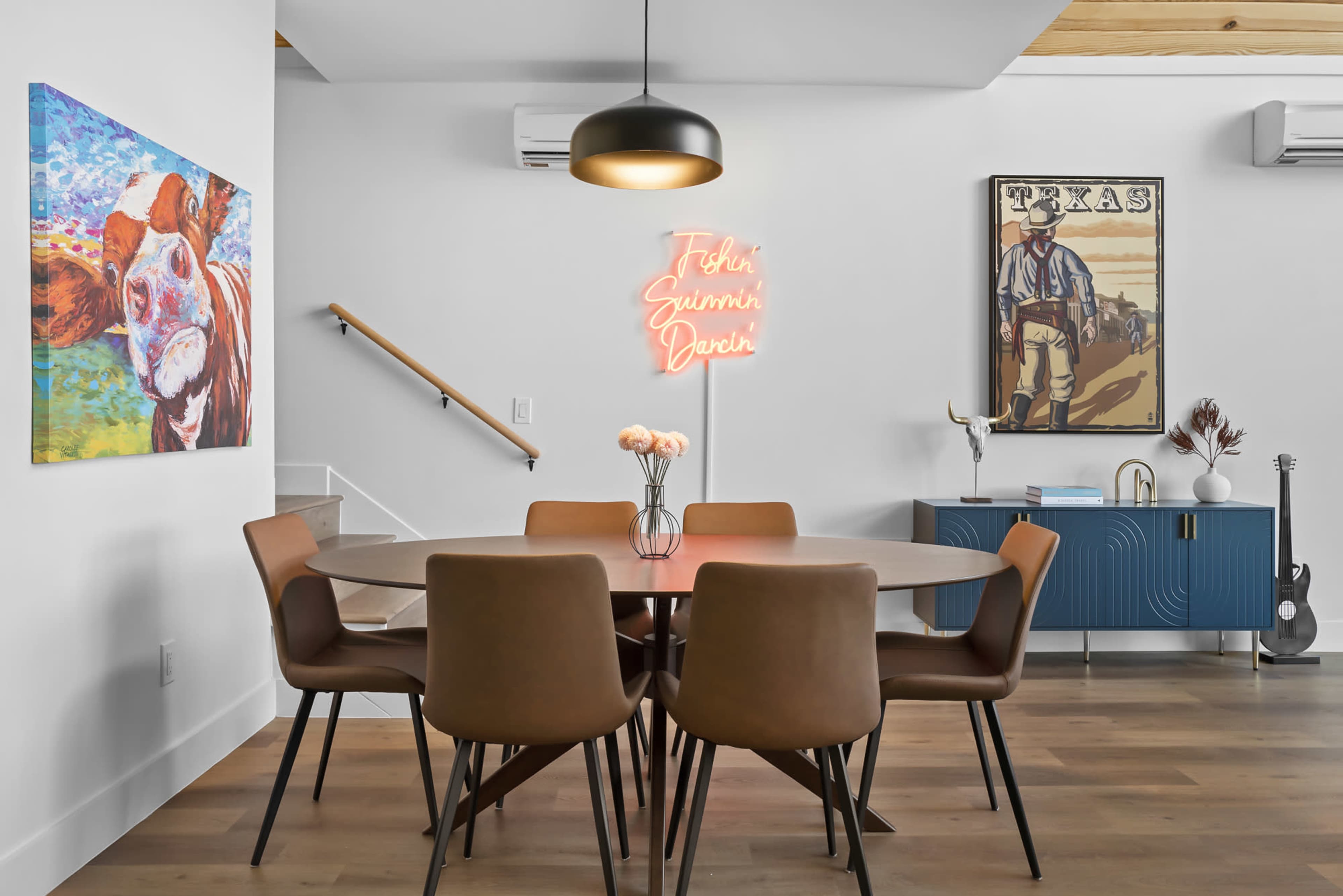 A modern dining area with a round table surrounded by six brown chairs, a decorative wall with art, and a neon sign.
