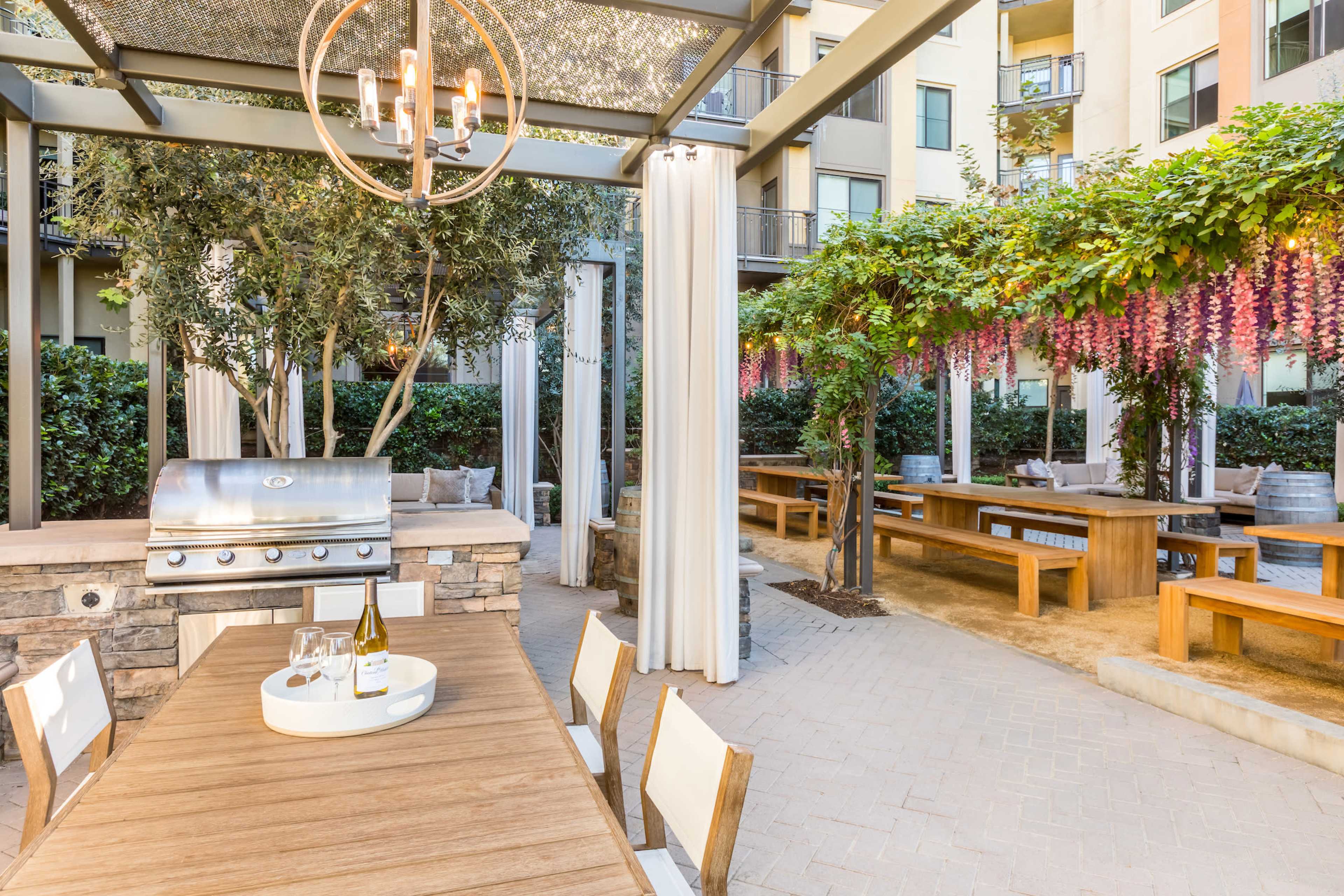An outdoor grilling area with a stainless steel BBQ grill, wooden dining tables, and plants draped with purple flowers under a shaded pergola.