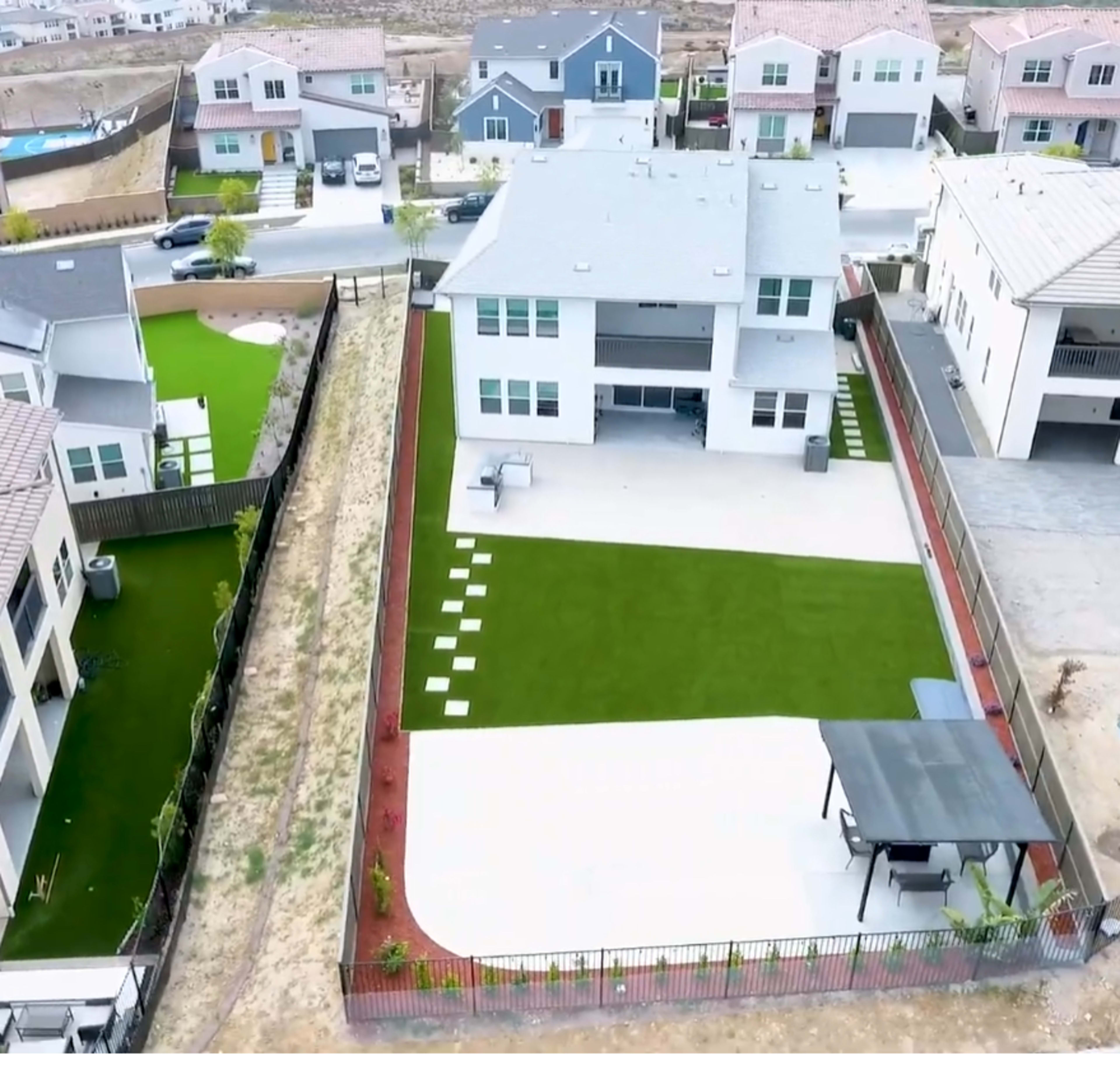 The image shows a residential neighborhood with several houses, featuring a yard that includes artificial grass, a patio area, and a gazebo.