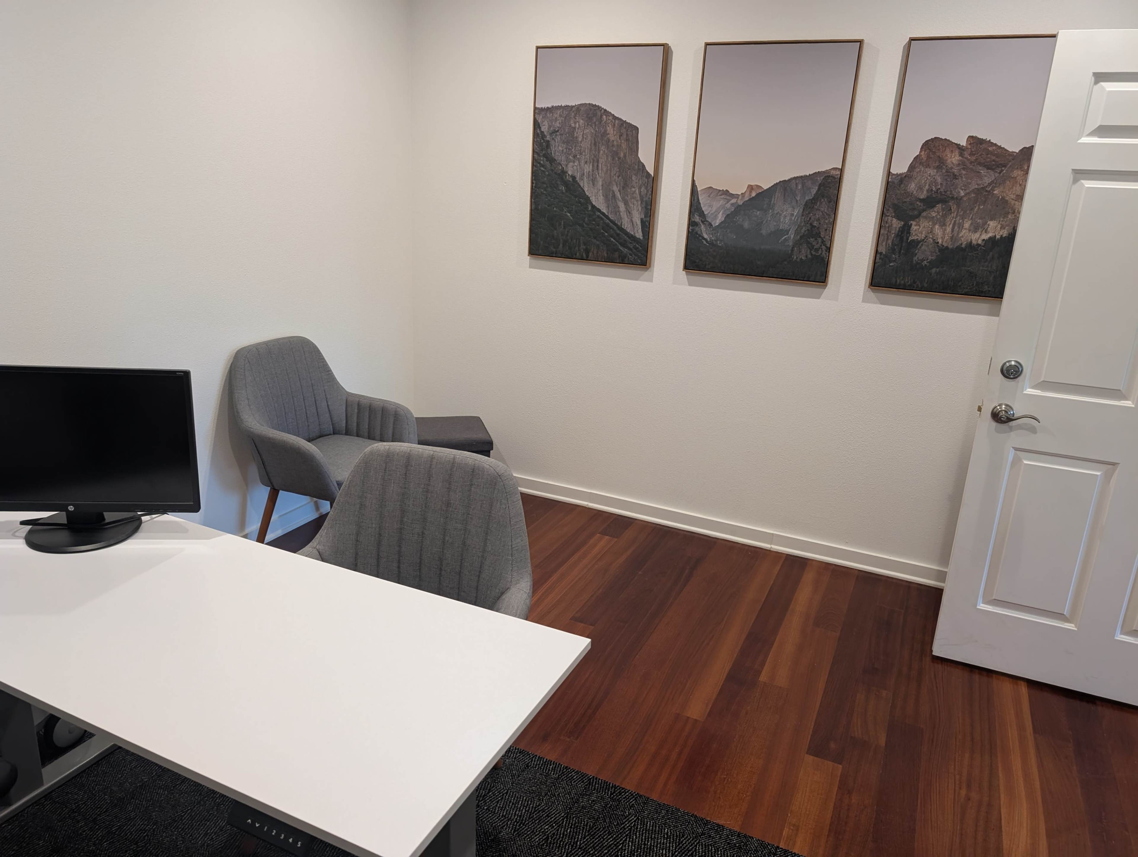 The image shows a minimalist office space with a white desk and monitor, two gray chairs, and three mountain landscape paintings on the wall.