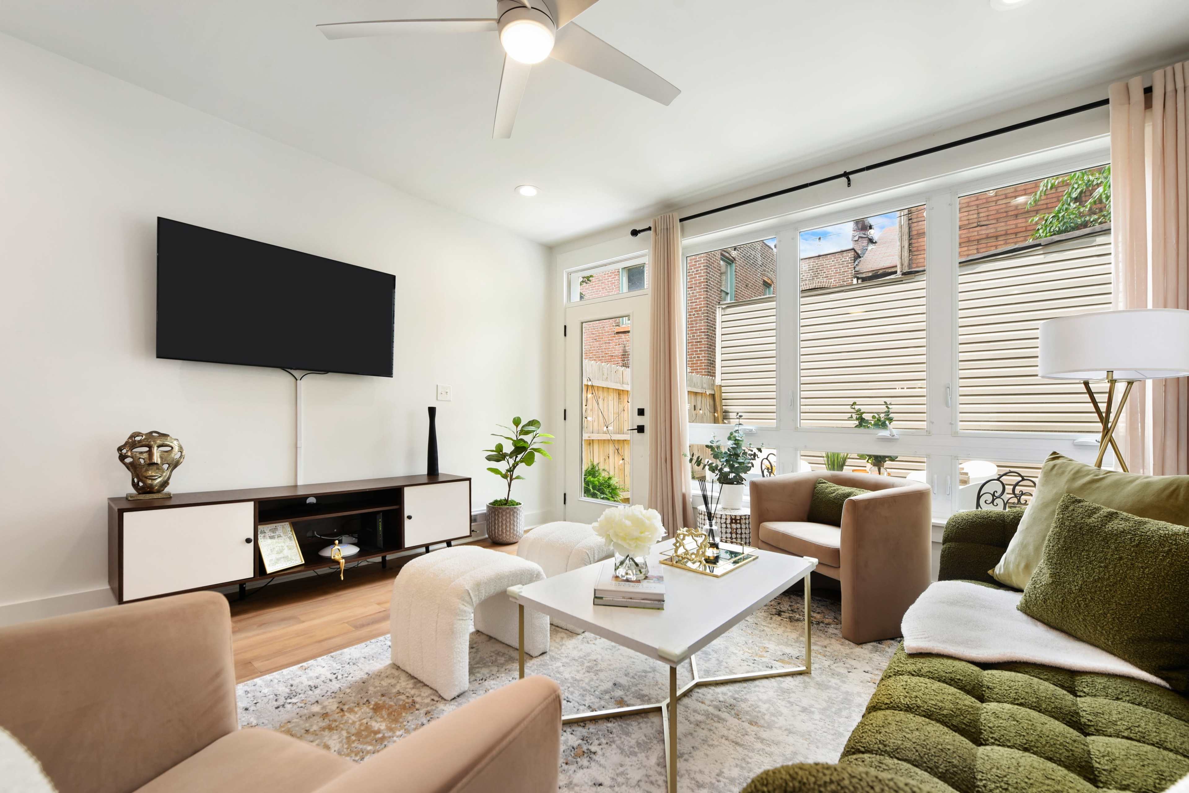 The image shows a modern living room with a green sofa, a beige armchair, a coffee table, and a wall-mounted television, featuring large windows that provide natural light and a view of an outdoor area.
