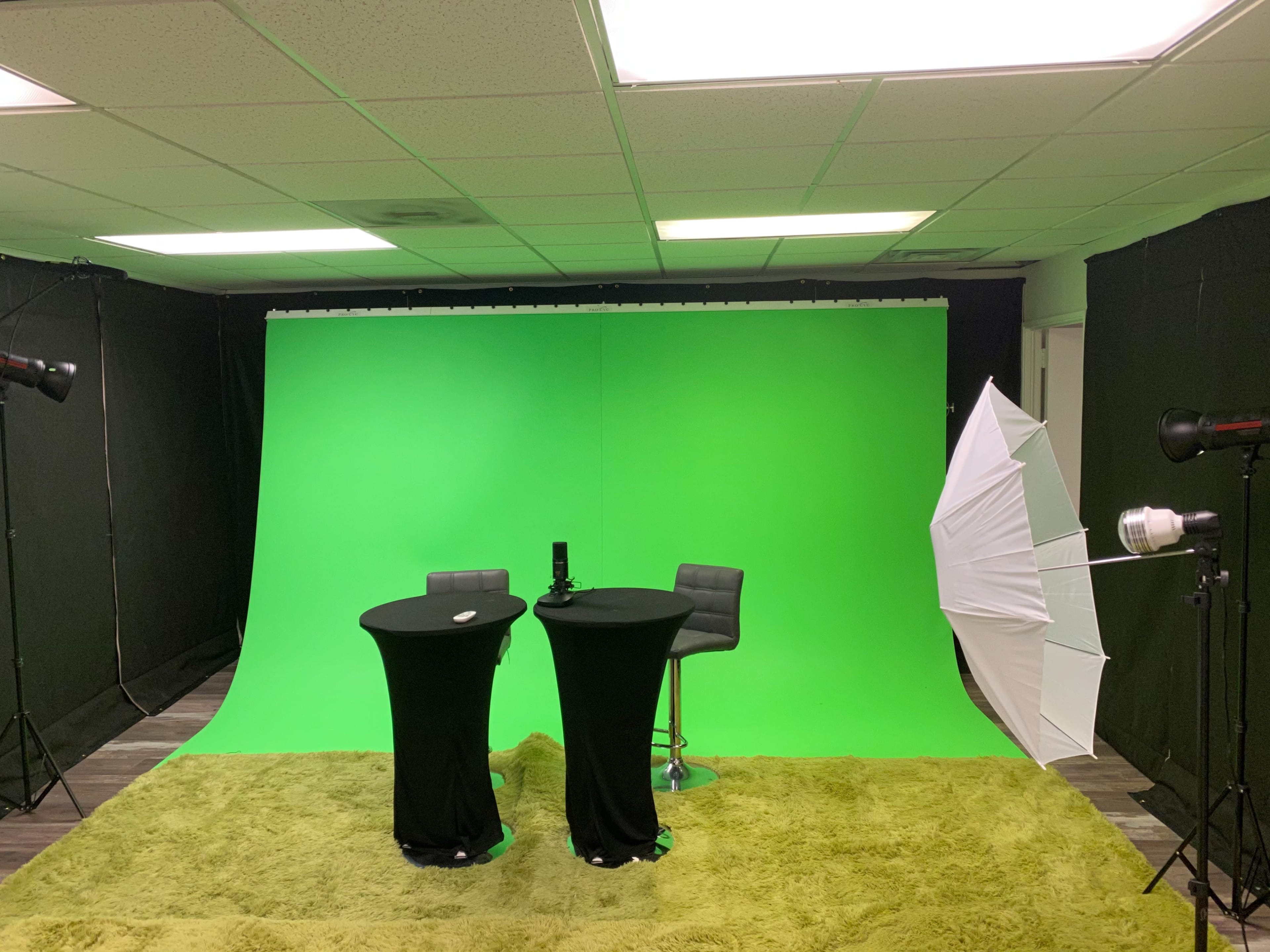 A studio setup featuring two black tables, a microphone, and lighting equipment in front of a green backdrop.