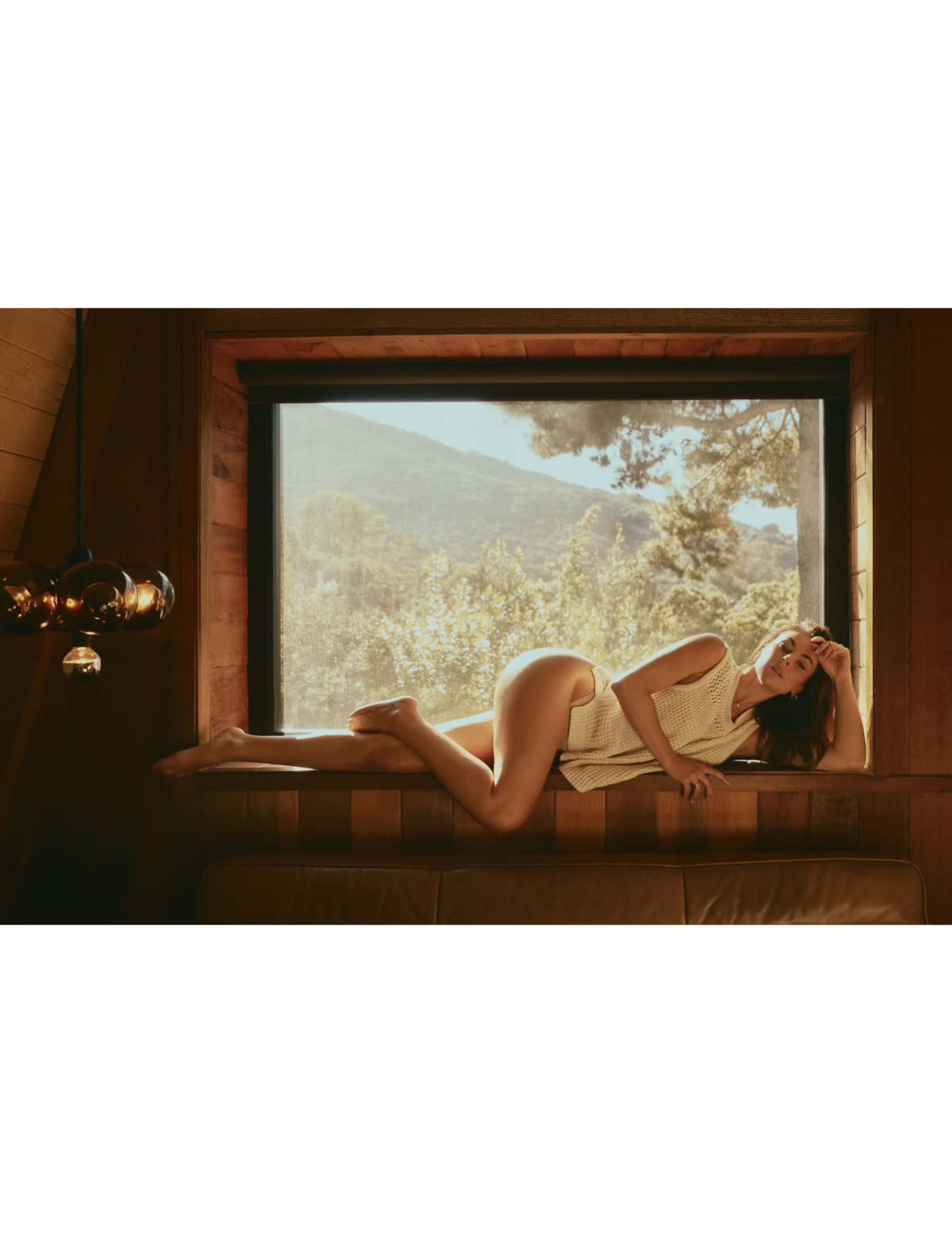 A person reclines against a large window, framed by a wooden interior, with a view of mountains and trees in the background.