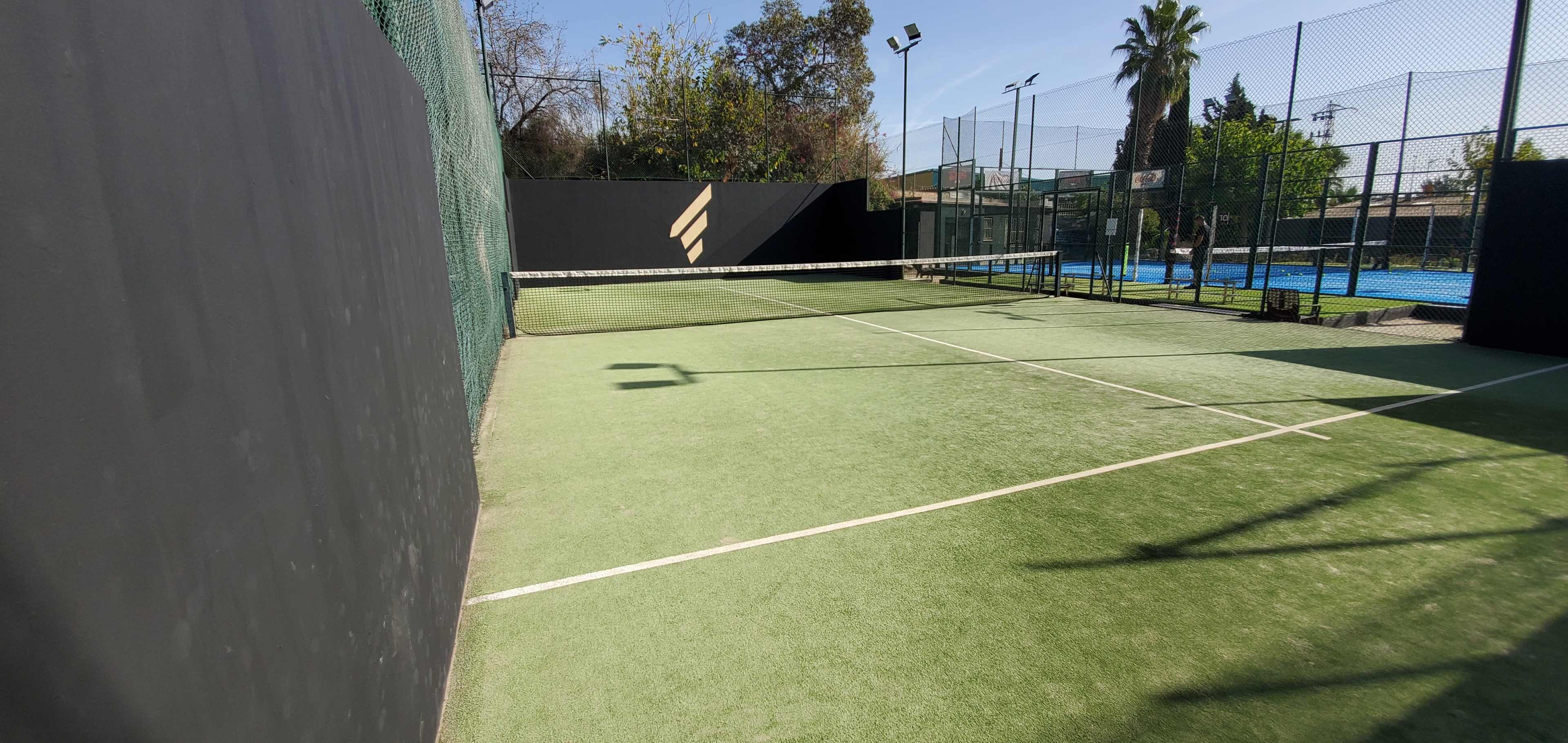 A tennis court with a green surface and a black fence is visible, featuring a net in the center and neighboring fenced courts in the background.