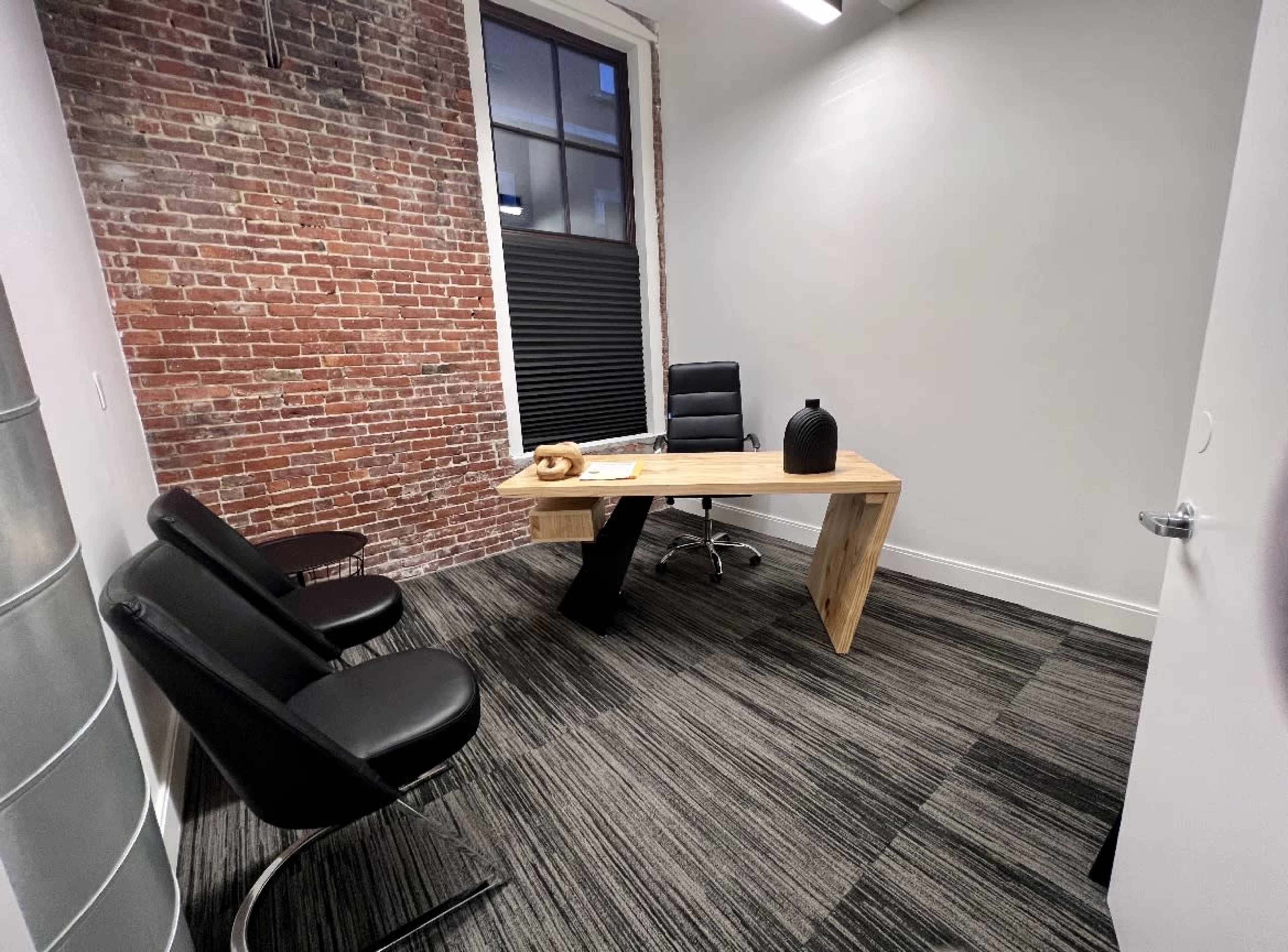 The image shows a small office with a wooden desk, a black chair, and two black lounge chairs against a brick wall.