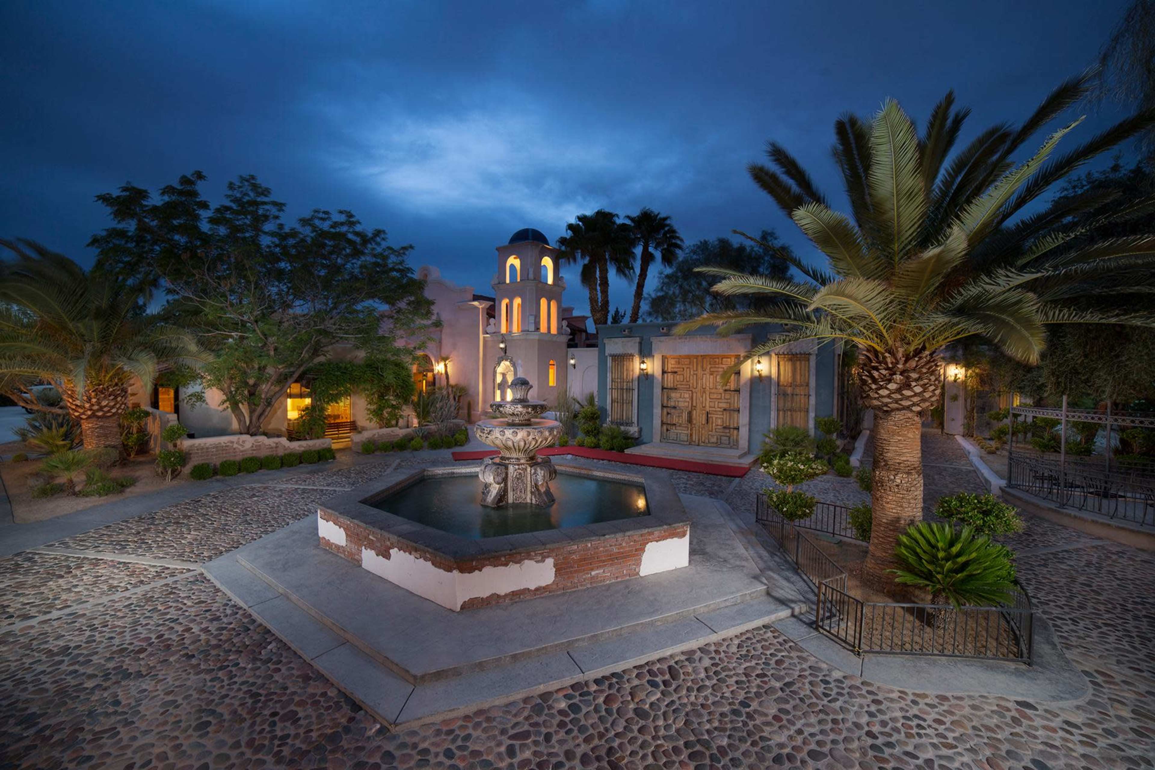 A decorative fountain is situated in a courtyard surrounded by palm trees and buildings under a twilight sky.