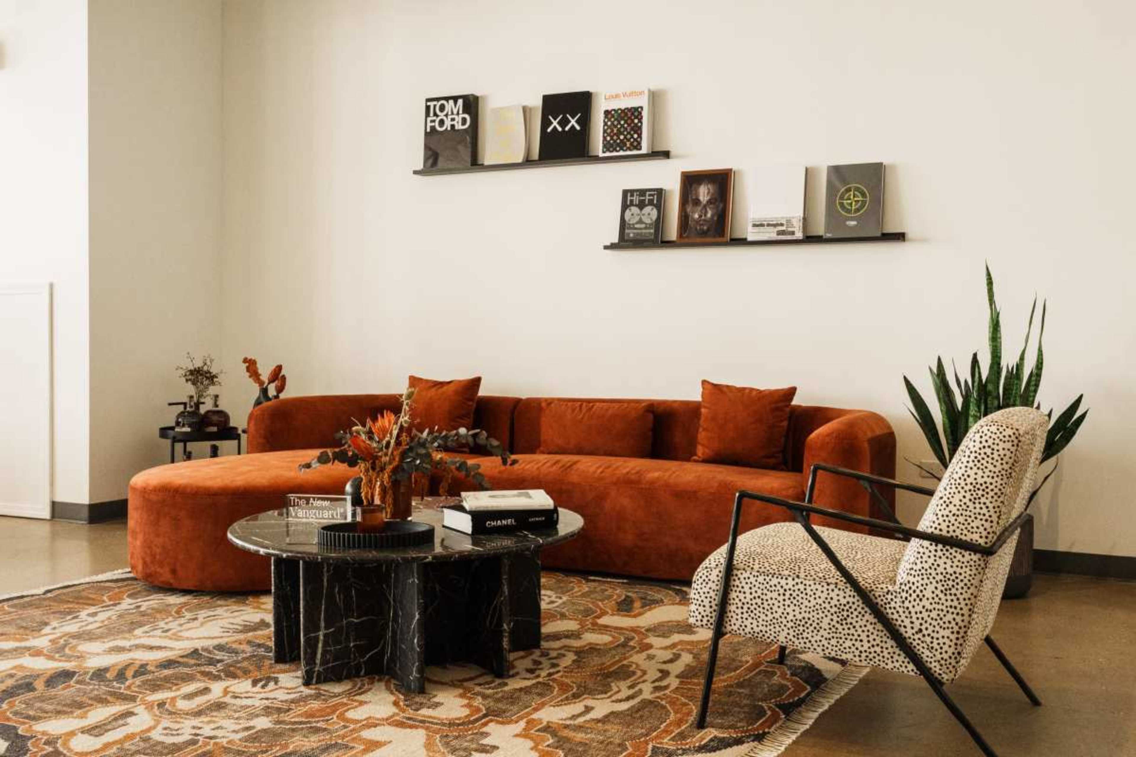 The image shows a modern living room featuring an orange curved sofa, a round black coffee table with decorative items, a patterned area rug, and a leopard-print accent chair.
