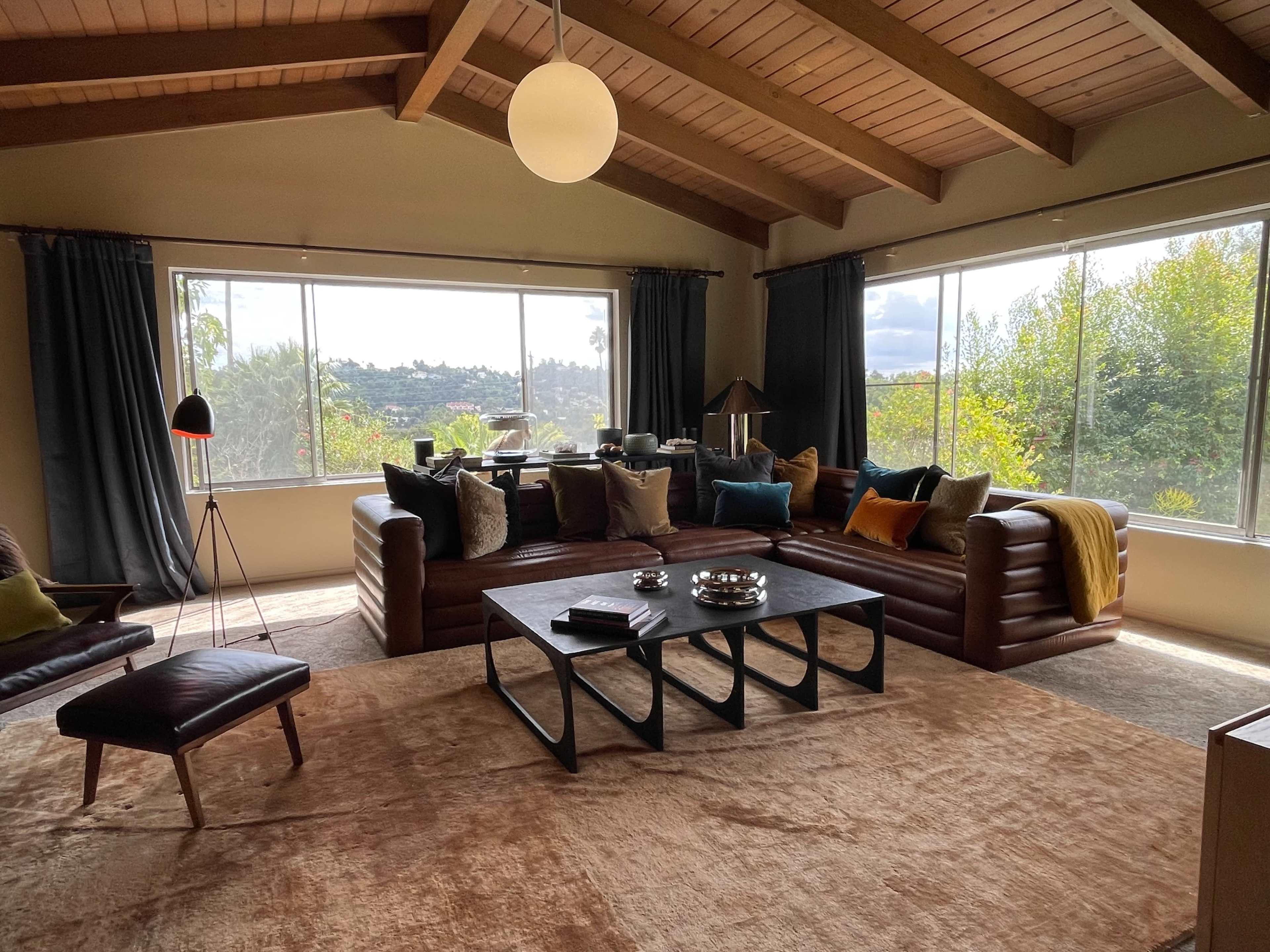 The living room features a large sectional sofa, a coffee table with decorative items, and large windows showcasing a view of trees and hills.