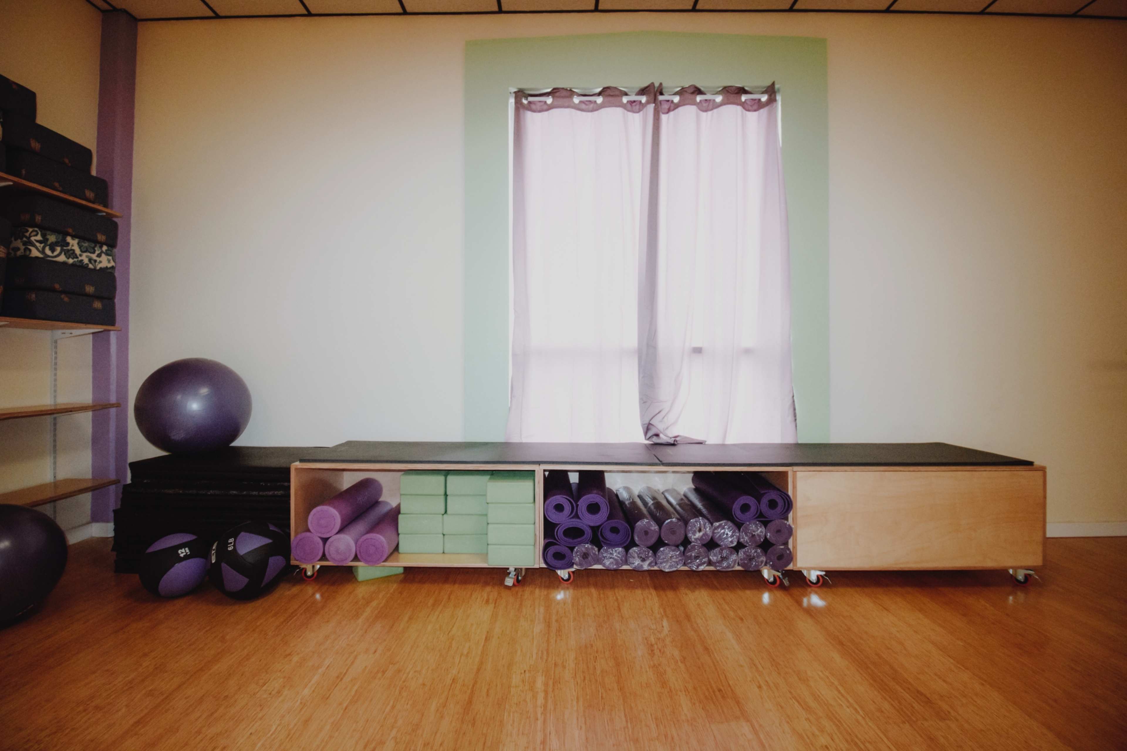 A studio space with exercise mats, a stability ball, and neatly arranged storage for yoga equipment beside a window with light purple curtains.