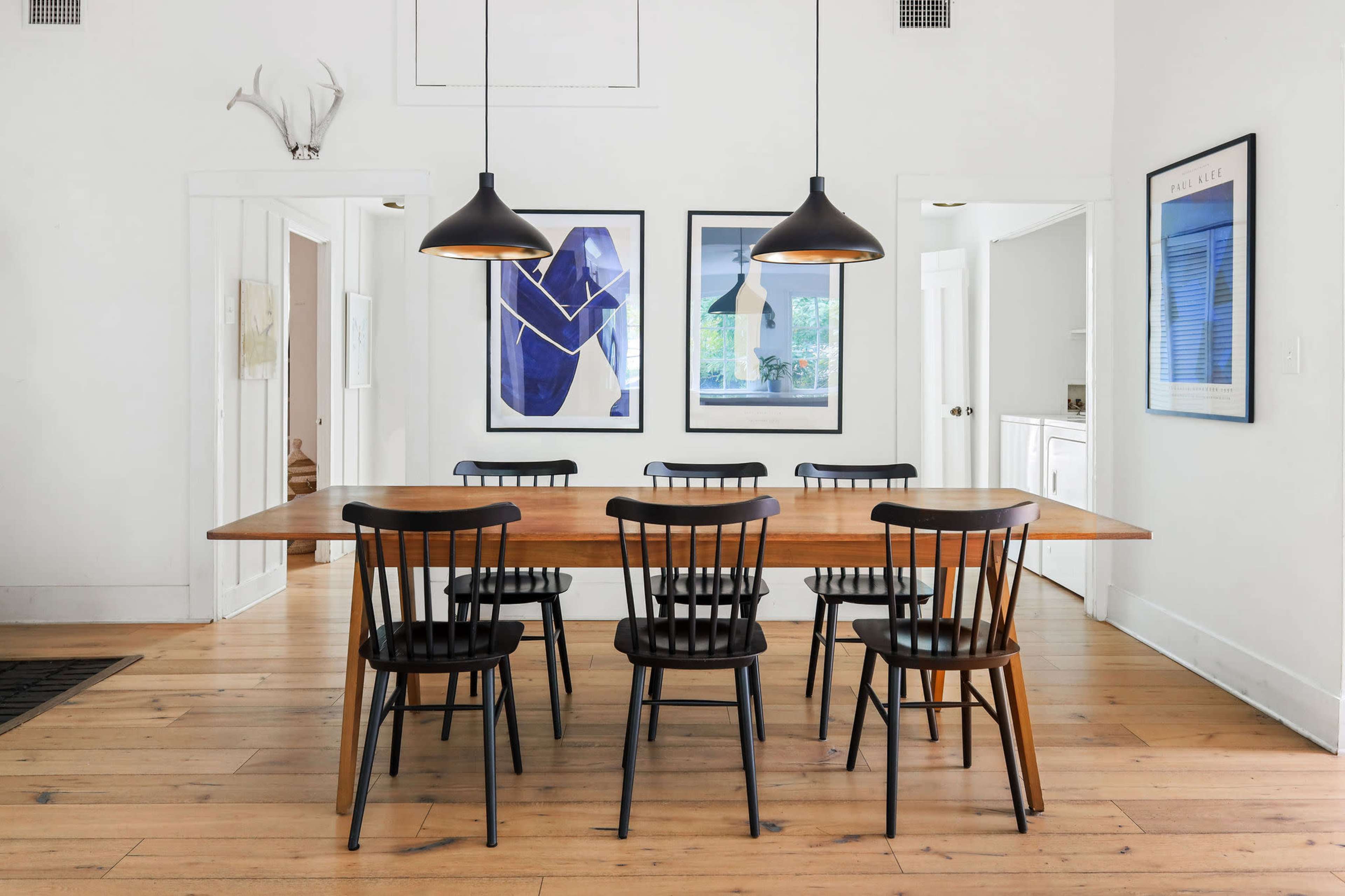A dining area features a wooden table surrounded by six black chairs, with two framed blue abstract artworks hanging on the white walls.
