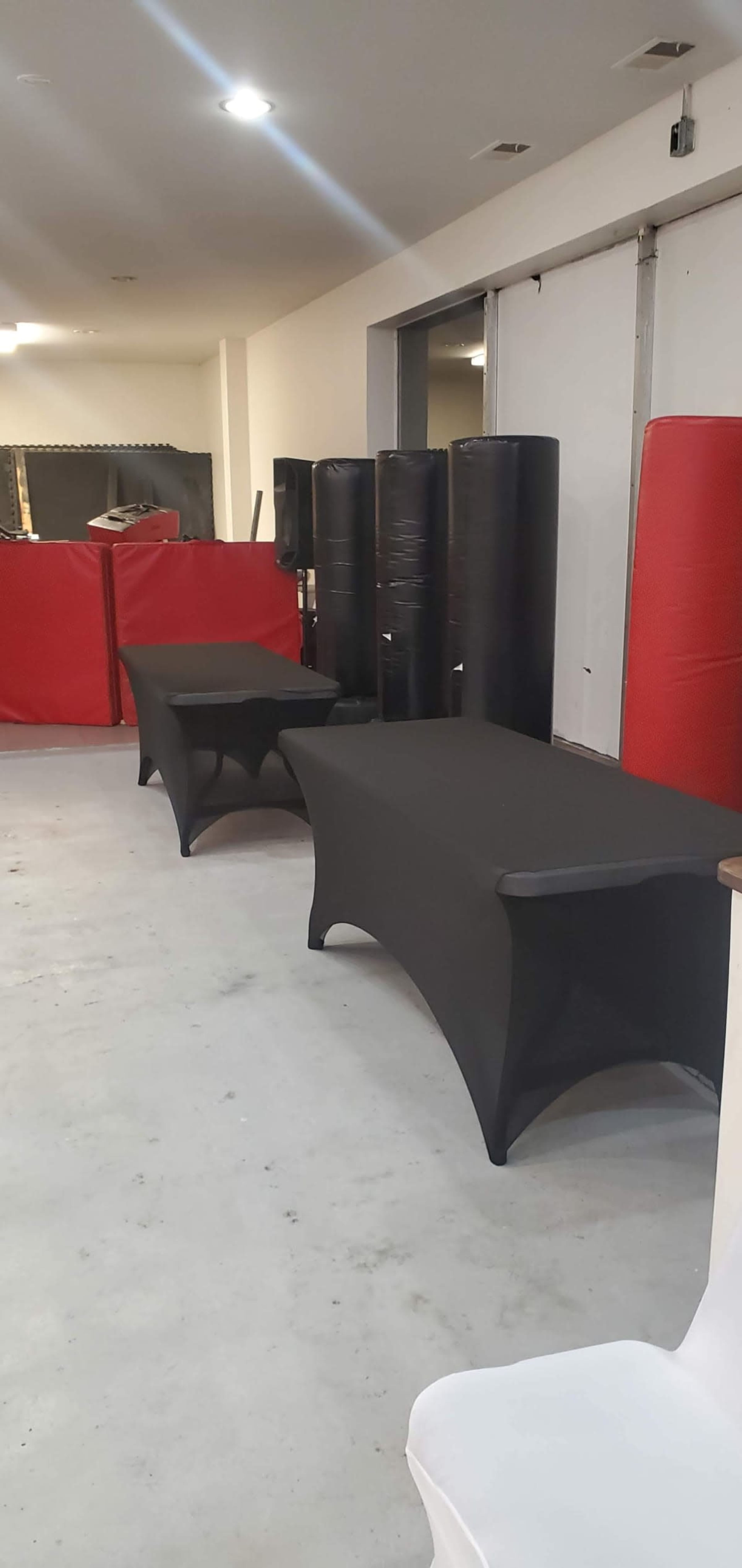The image shows a room with two black fitted tables and various pieces of gym equipment in the background, including red and black padded columns.
