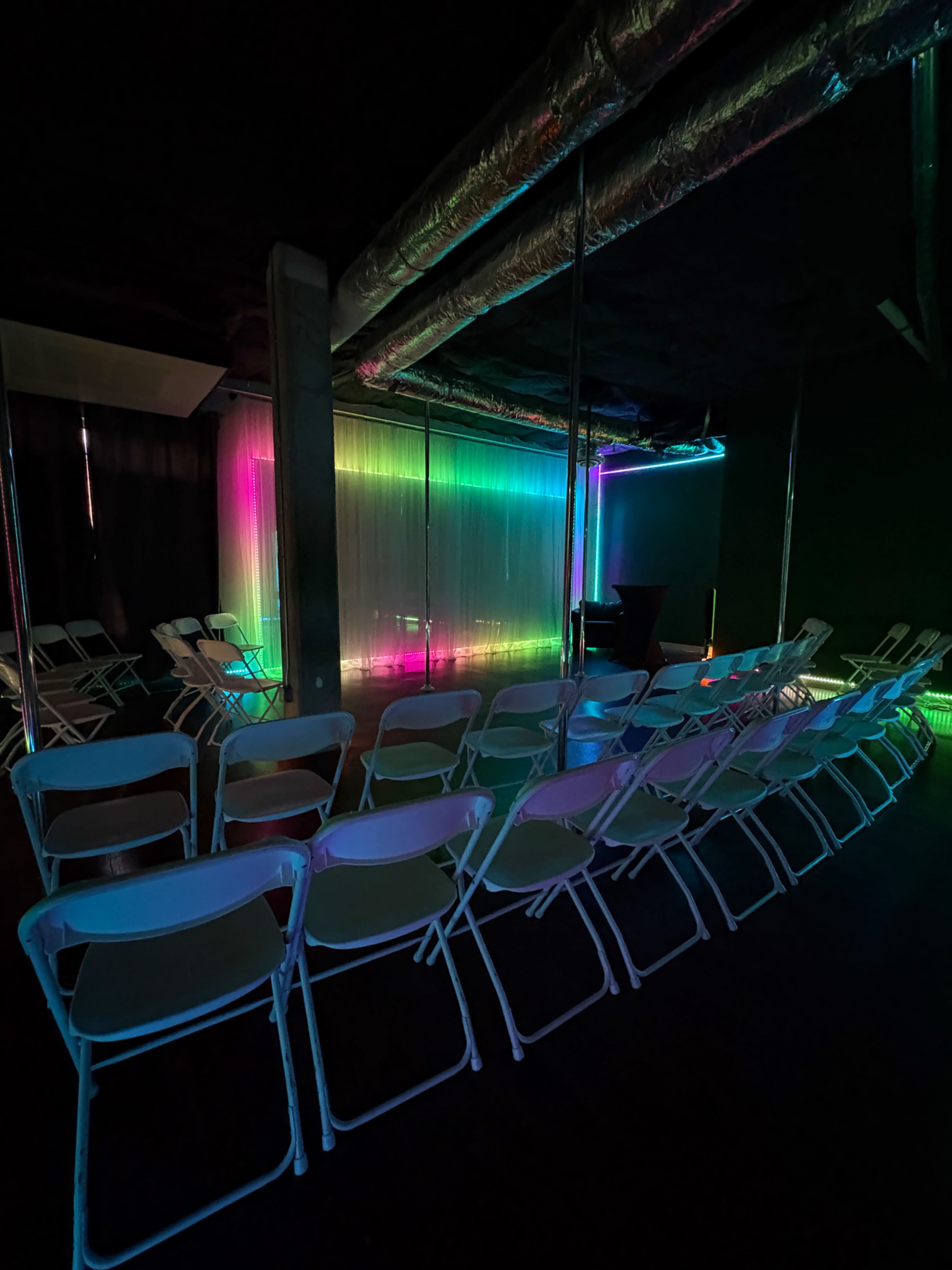 The image shows an empty room with rows of white folding chairs facing a brightly lit backdrop with colorful neon lighting.