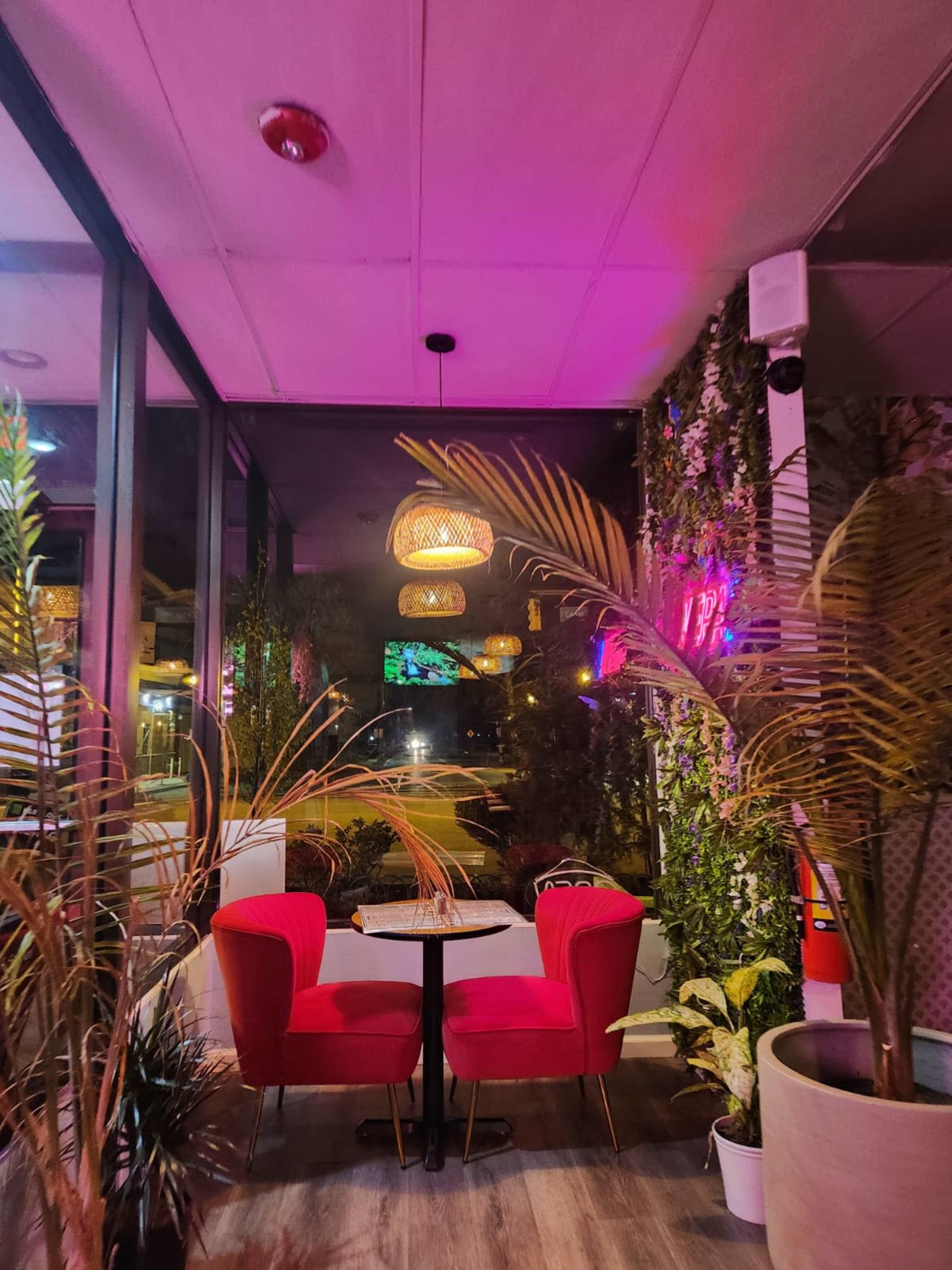 A cozy café corner features two red chairs and a small table, surrounded by plants and illuminated by colorful lighting.
