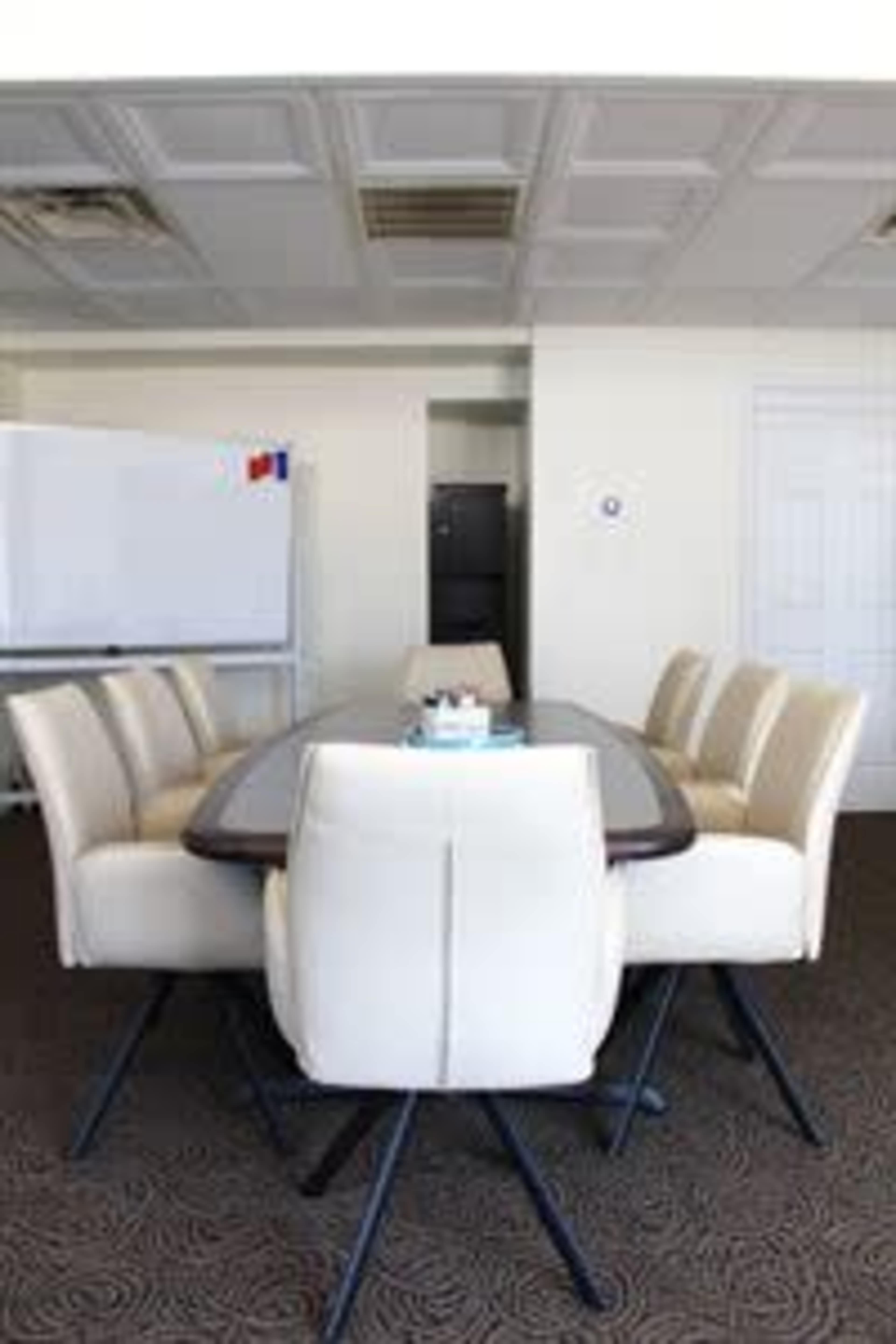 A conference room features a large oval table surrounded by cream-colored chairs, with a whiteboard and kitchen area visible in the background.