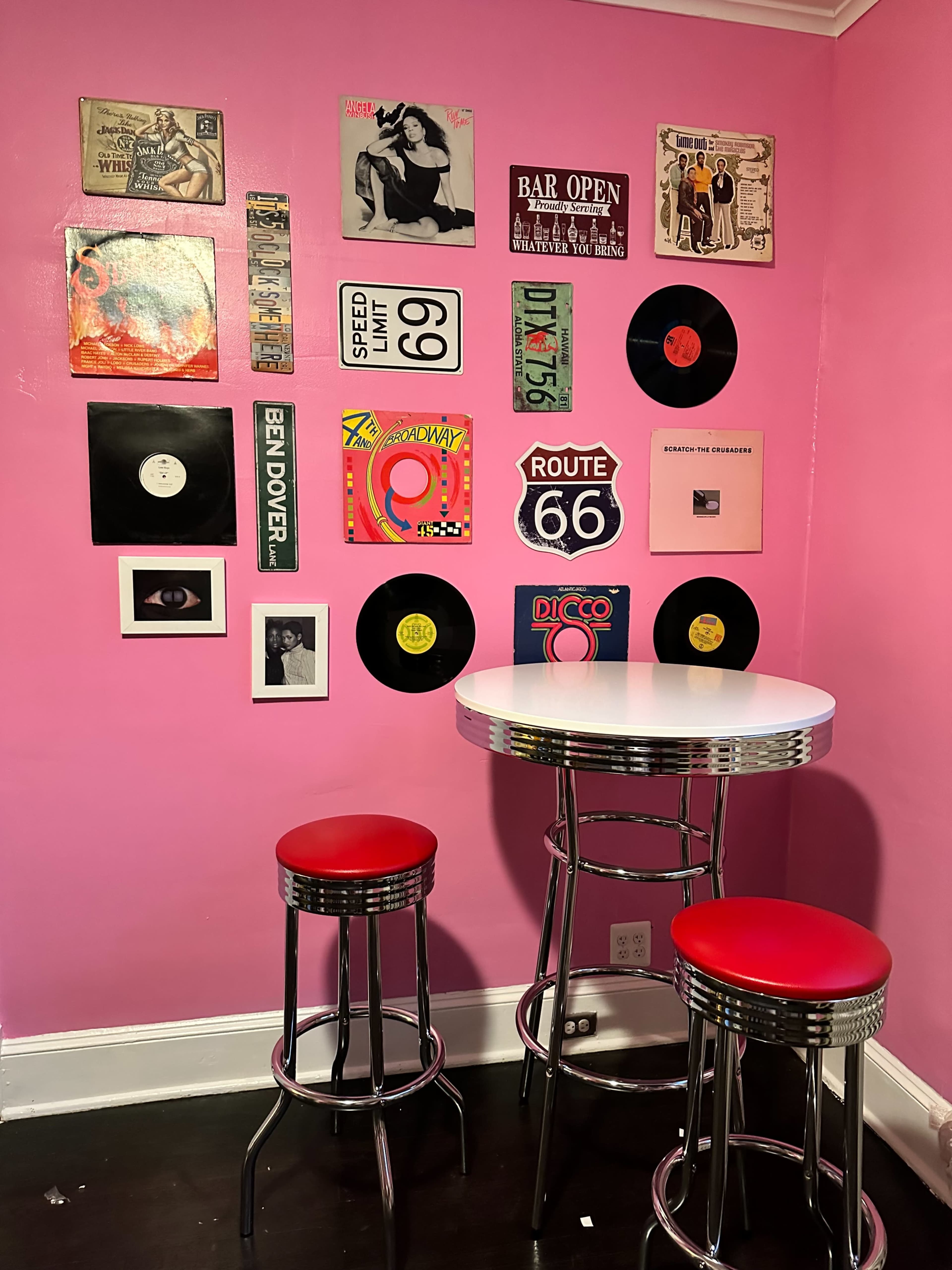 A vibrant pink wall adorned with retro music-themed art and vinyl records, with a small circular table and two red barstools in front.