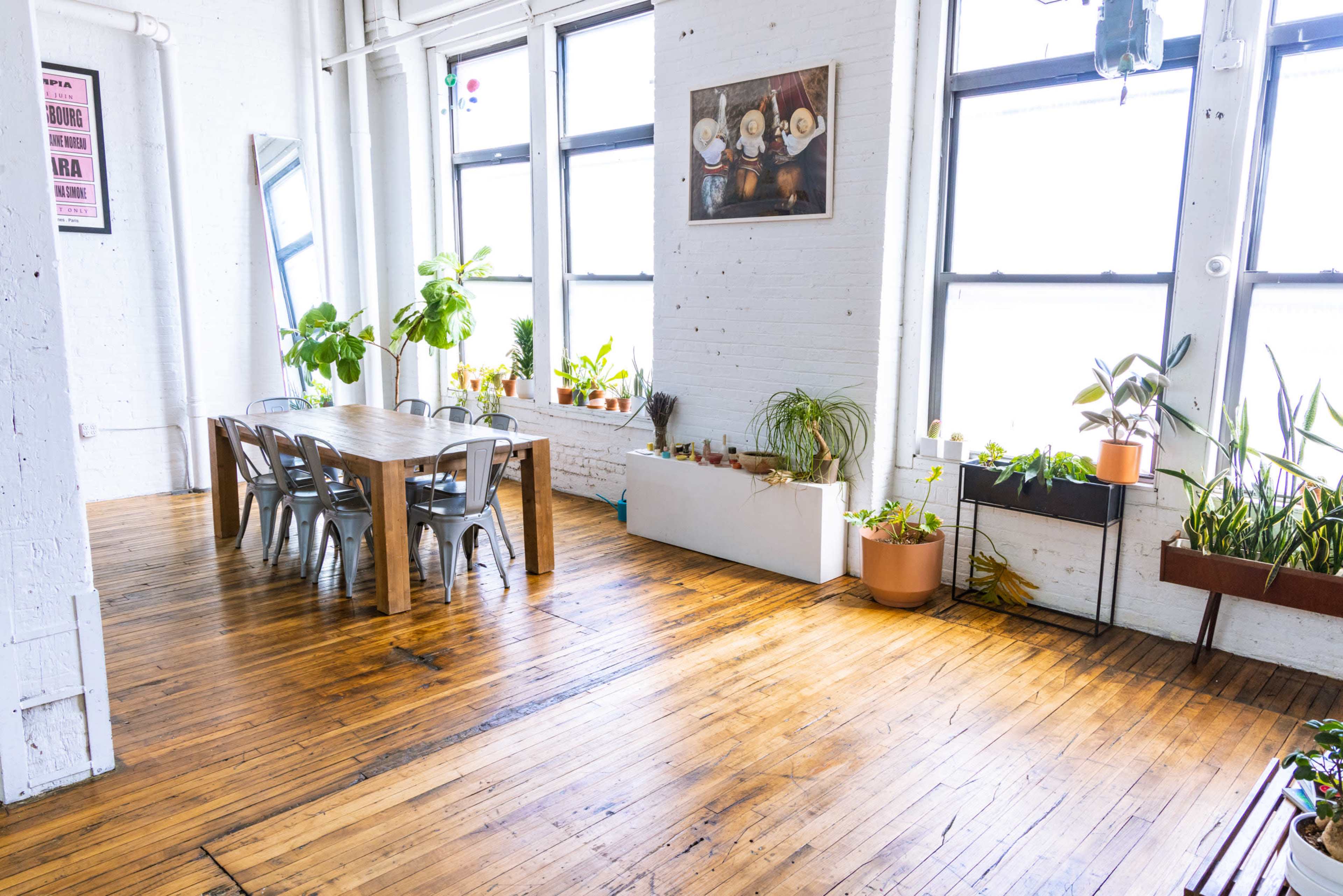A spacious room features a wooden dining table surrounded by metal chairs, large windows, and various potted plants along the walls.