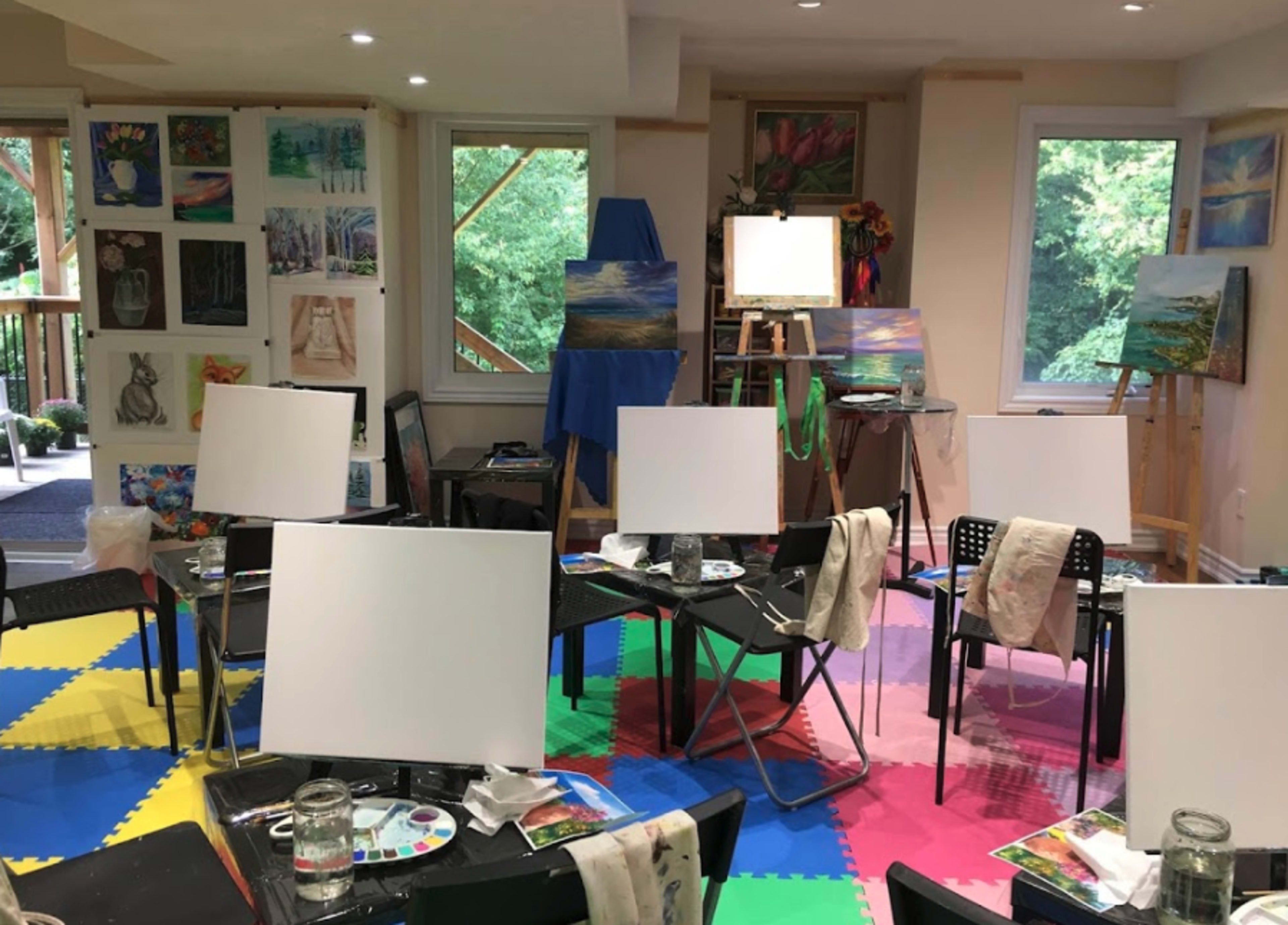 An art studio filled with empty canvases on black easels, surrounded by colorful painted walls and art supplies on tables.
