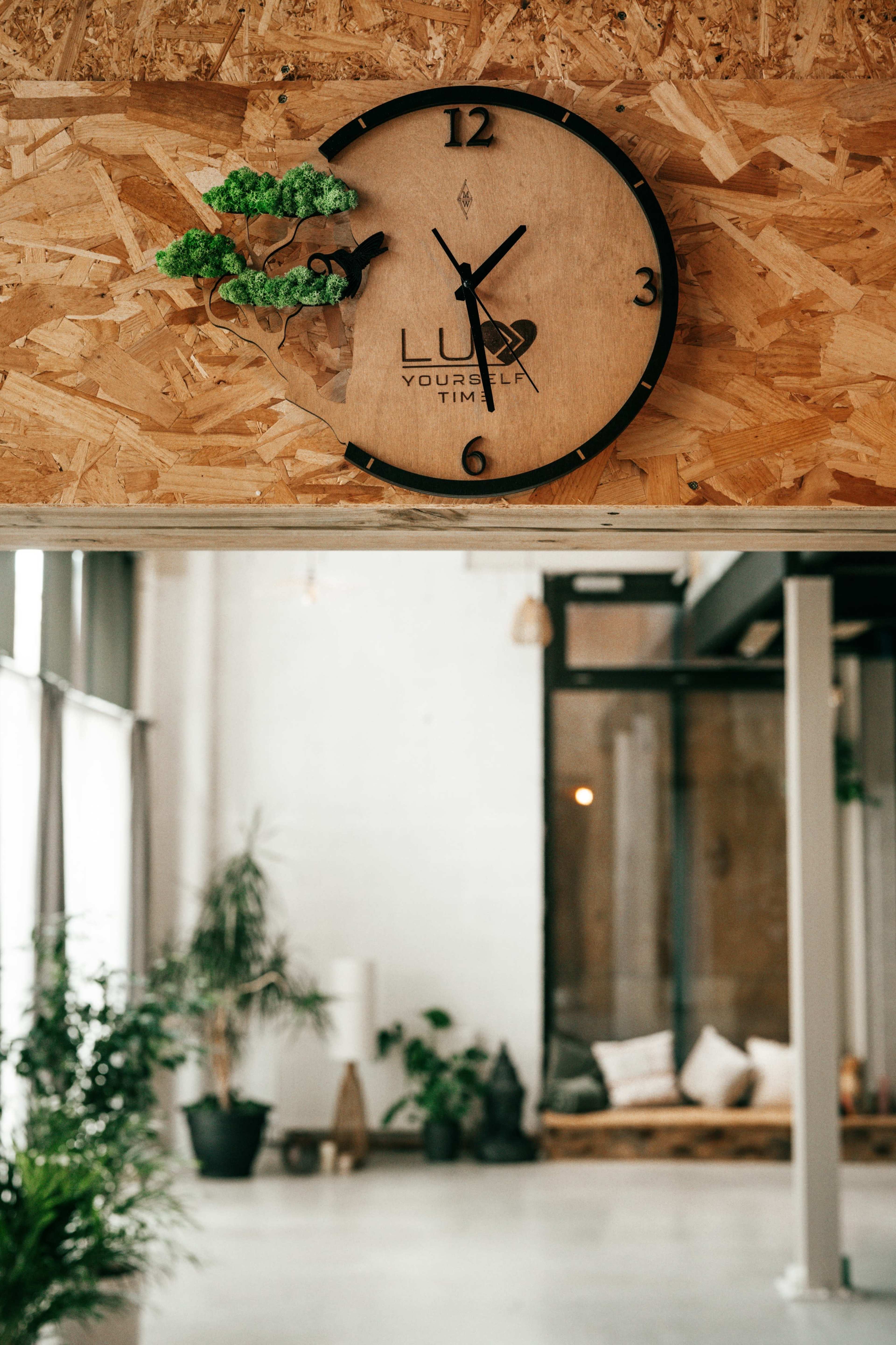 A wall clock with a wooden finish and green accents is mounted on an intricately designed wooden panel in a spacious, modern interior.
