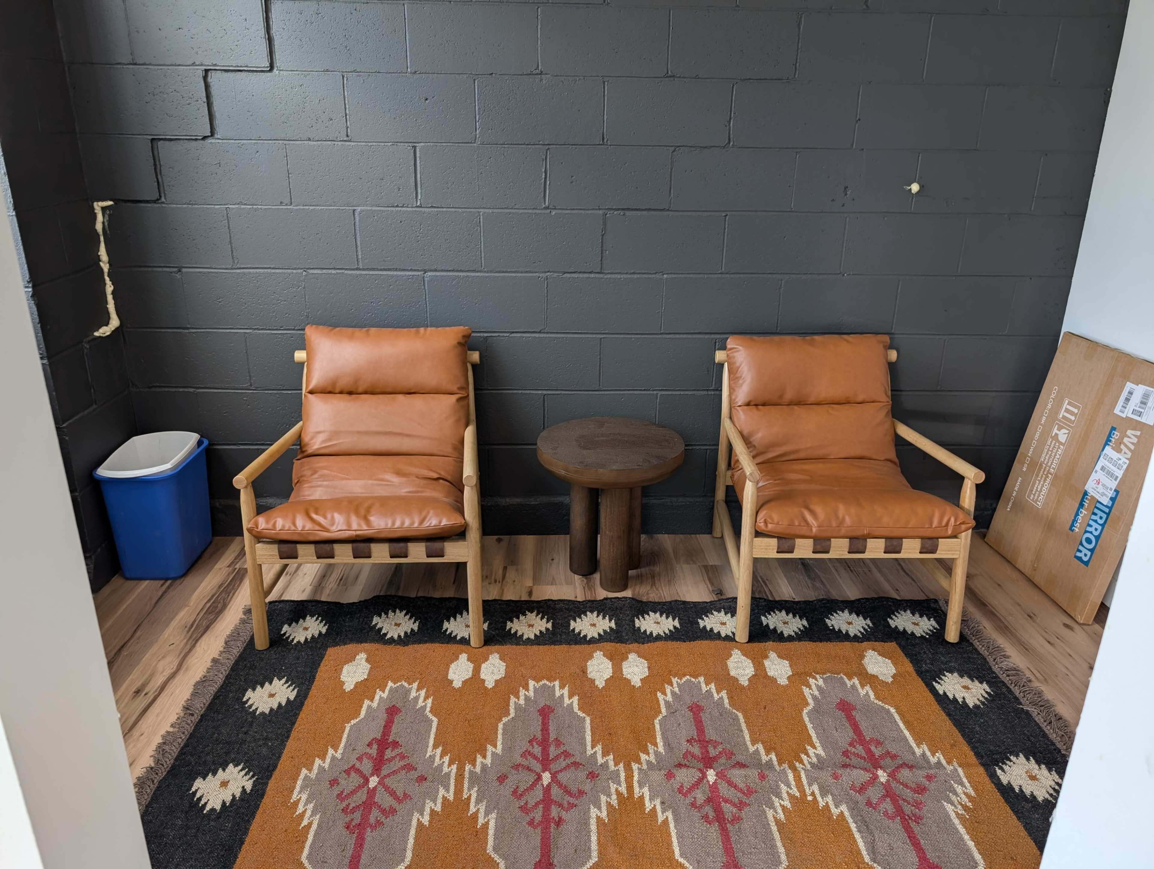 The image shows a small room with two leather armchairs facing a round wooden table, a blue recycling bin in the corner, and a patterned rug on the wooden floor.