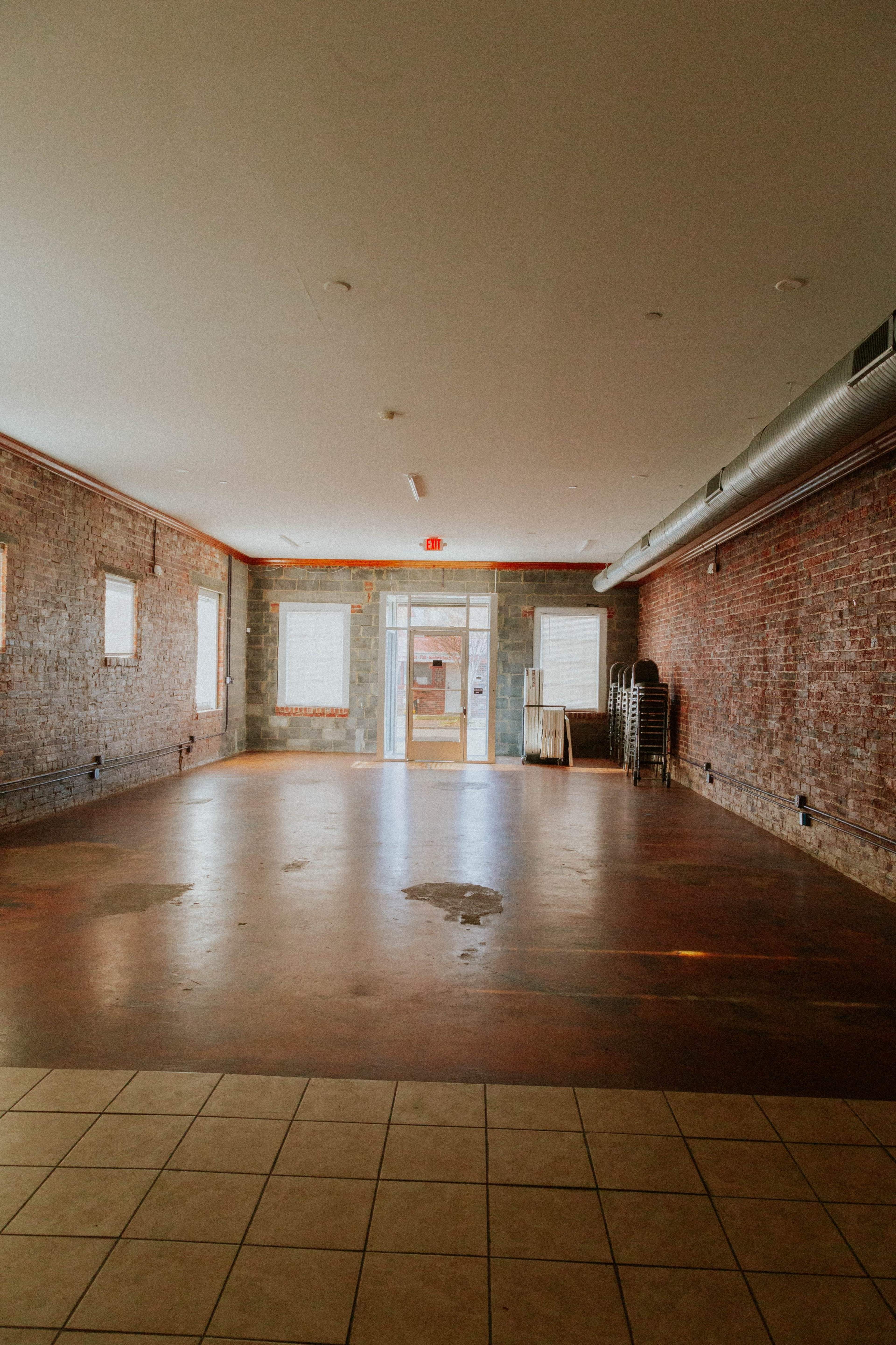 The image shows a spacious, empty room with brick walls, a tile floor, and a doorway leading outside.