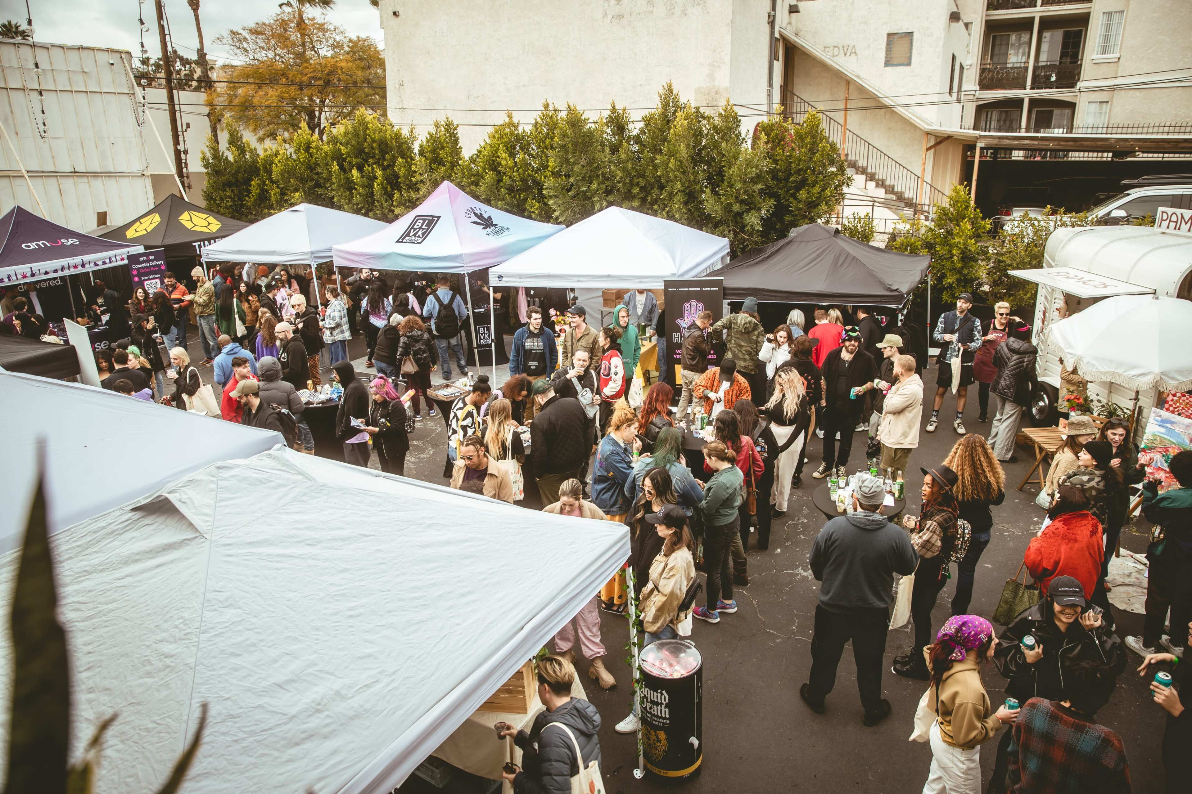 A large crowd gathers in a parking lot filled with various tents, with people socializing and vendors displaying their goods.